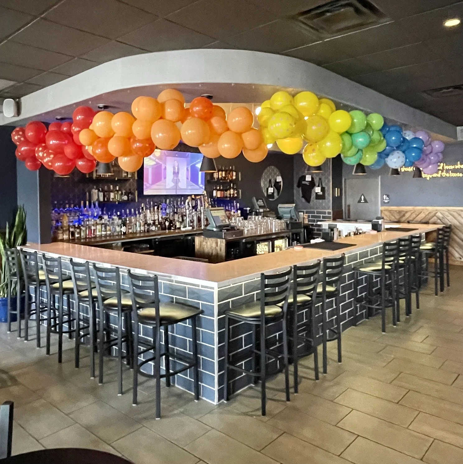 Bar with rainbow-colored balloons hanging from the ceiling, bar stools along the counter, and a TV screen above the shelves of bottles.