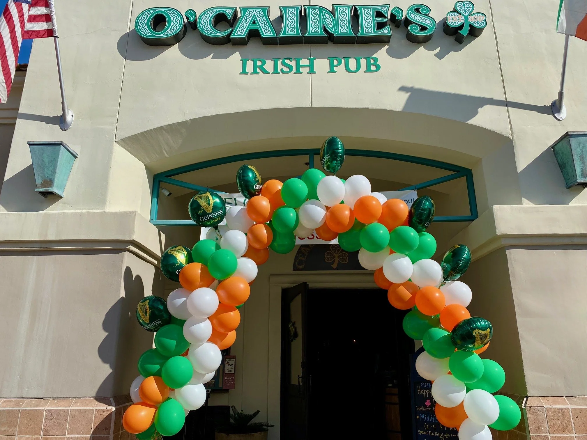 Entrance of O'Cainie's Irish Pub in Palm Springs, CA decorated with green, white, and orange balloons, and Union Jack balloons; pub sign with Celtic design, an American flag on the side.