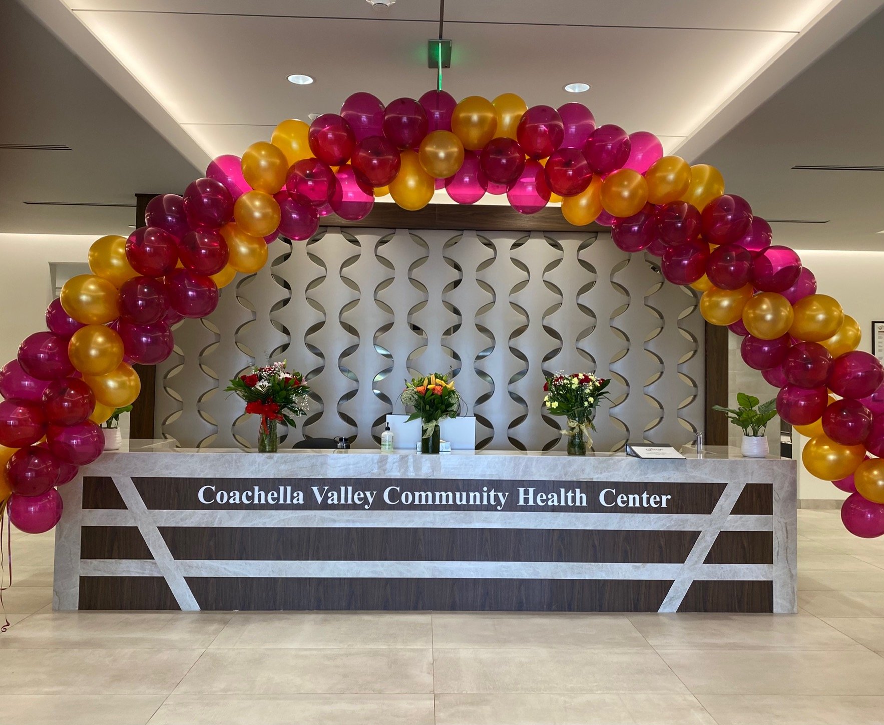 Decorative balloon arch in pink and yellow colors over the reception desk of the Coachella Valley Community Health Center, with flower arrangements on the desk and a patterned wall in the background.