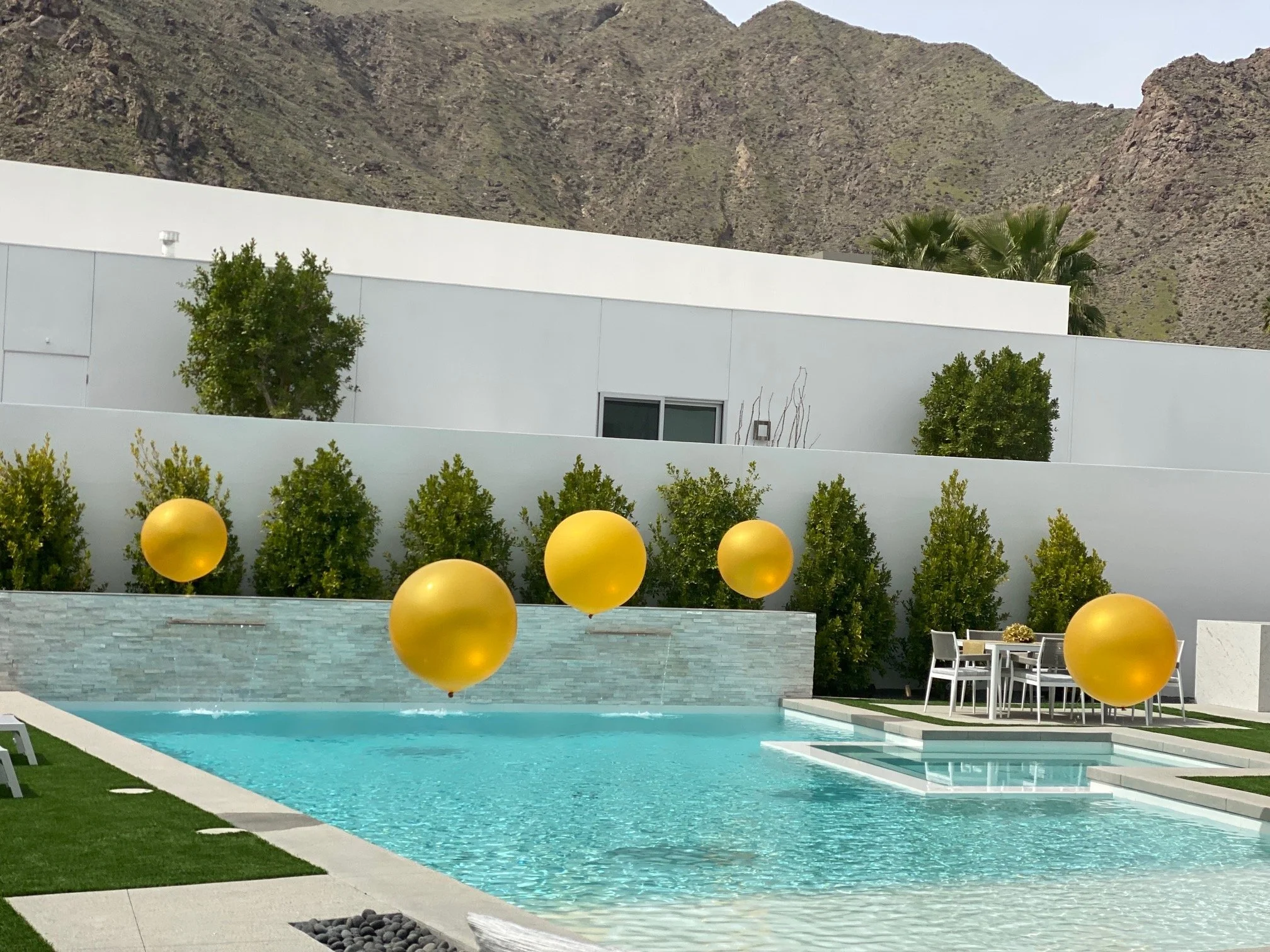 Modern outdoor pool area with turquoise water, surrounded by green grass and potted plants, with yellow balloons and outdoor seating, set against a backdrop of a white building and mountainous landscape.