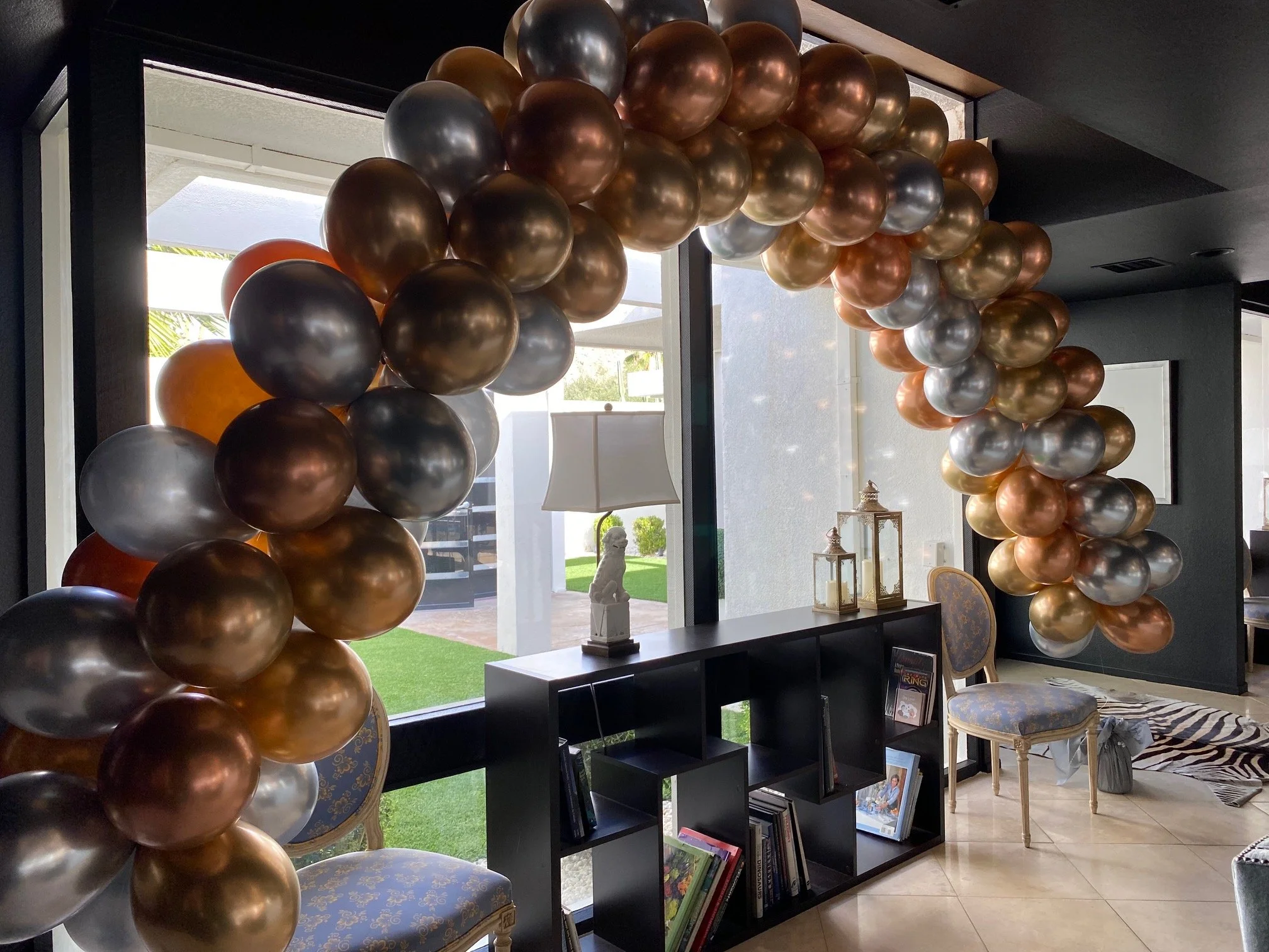 Indoor space decorated with metallic balloons in shades of gold, bronze, and silver, arranged around a window. Inside the room, there is a black bookshelf with framed pictures and books, a table lamp, a chair with patterned upholstery, and decorative