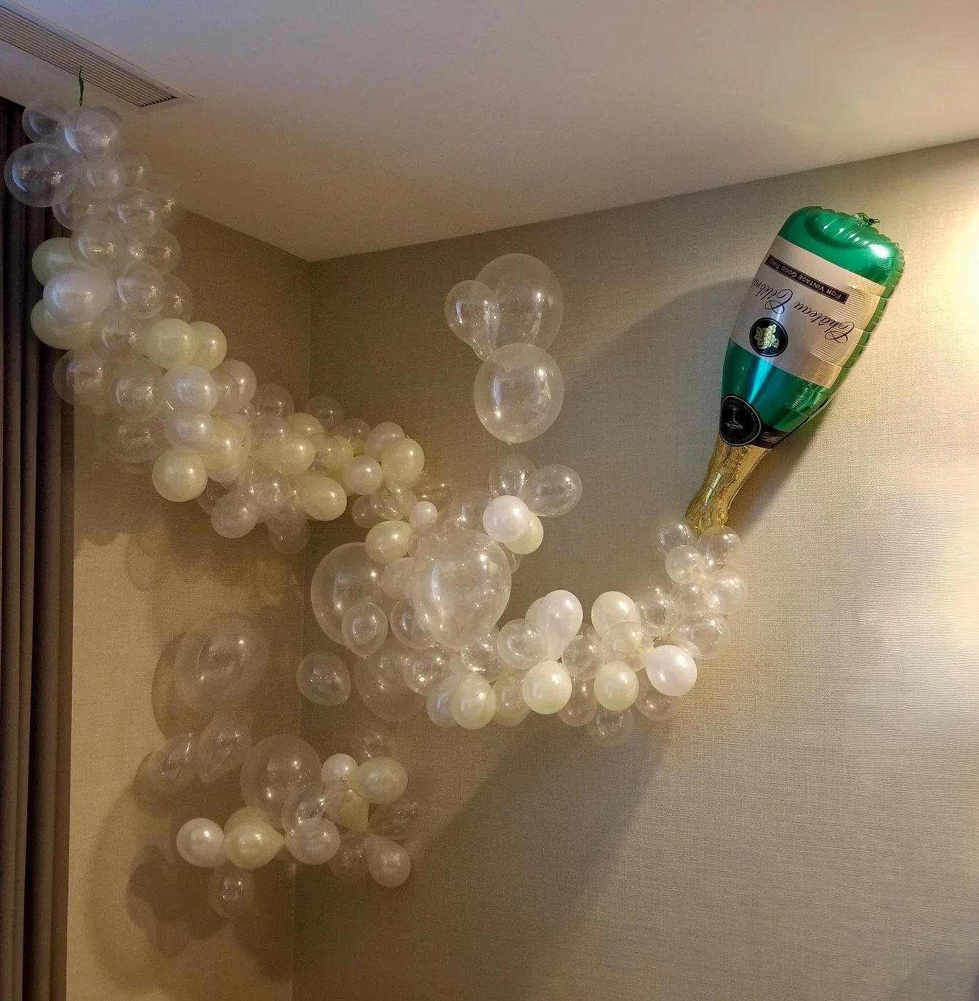 A cluster of white and transparent balloons arranged along a wall and ceiling, with a green and gold champagne bottle-shaped balloon on the right side.