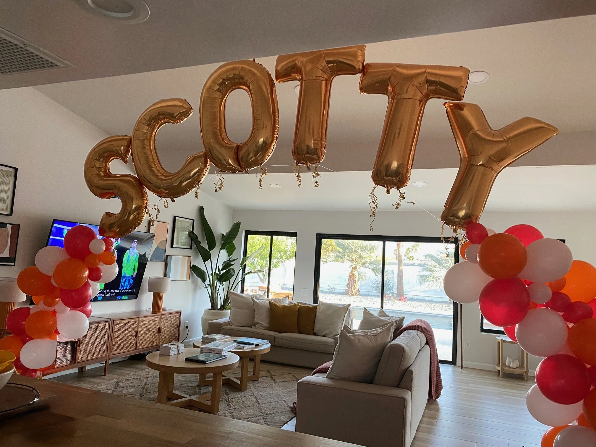 Living room decorated with orange, red, and white balloons forming columns and a large Gold 'SCOTTY' balloons hanging from the ceiling.