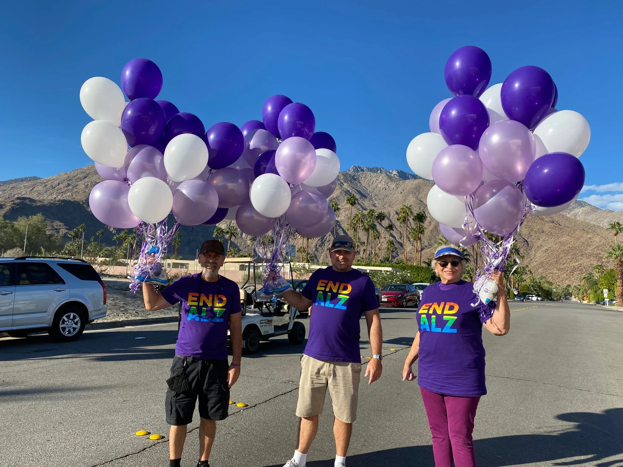 Three people standing in a parking lot holding bunches of purple and white balloons, with mountains and palm trees in the background.