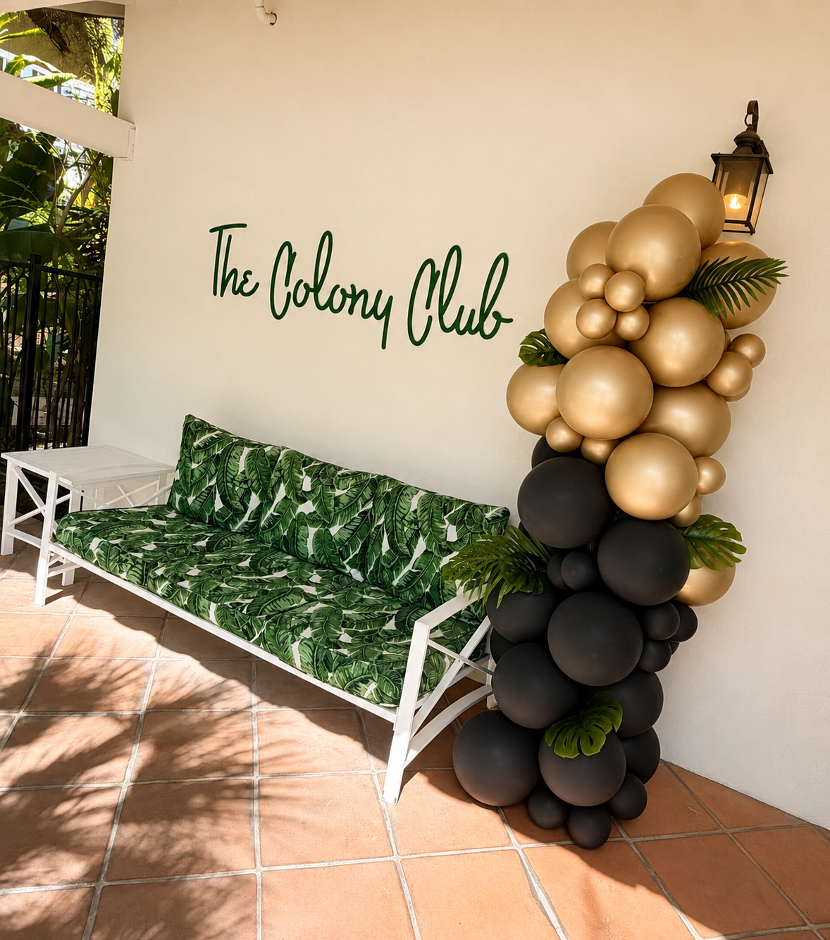 Decorative outdoor area with a white bench cushion with green tropical leaf pattern, a small white side table, a balloon arrangement with gold, black, and green balloons, and a sign reading 'The Colony Club' on the wall.