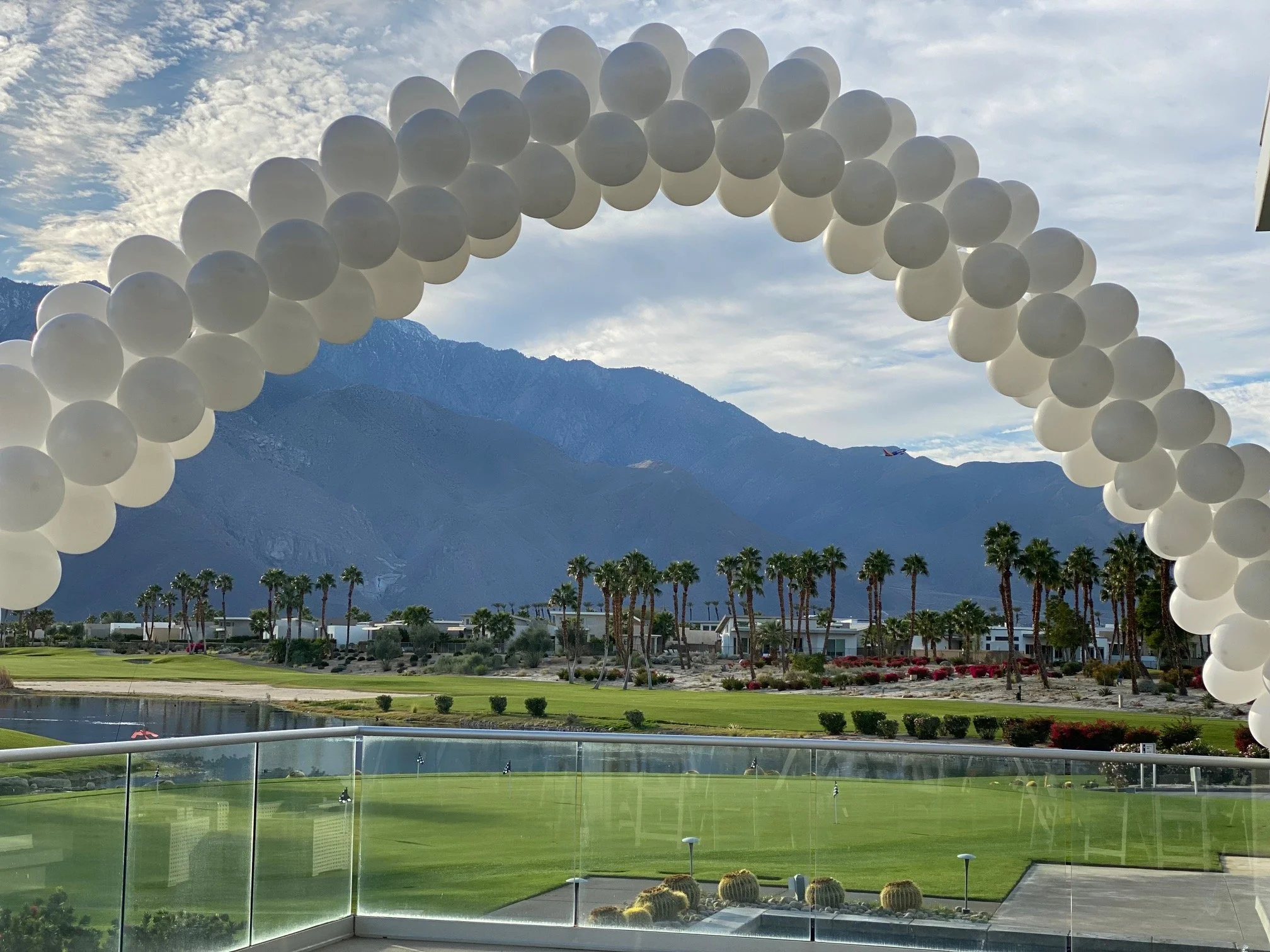 A cluster of white balloons arranged in an arch shape against a cloudy sky, with mountains and palm trees visible in the background.