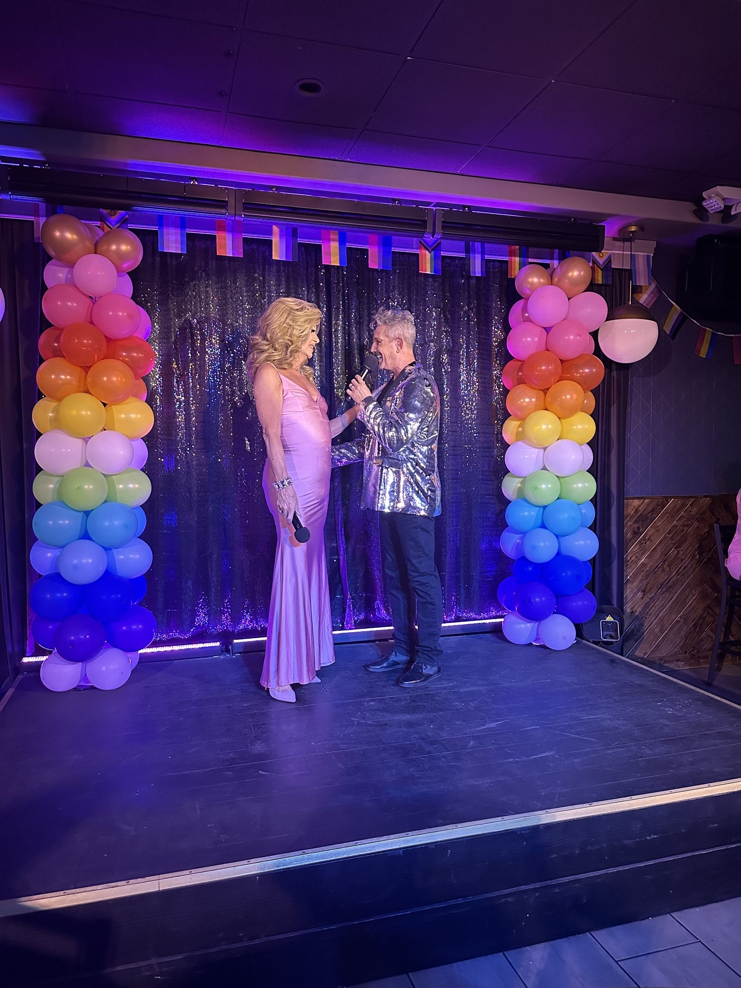 Two people in formal attire standing on a stage decorated with rainbow-colored balloon columns and a glittery black background, engaging in a conversation.
