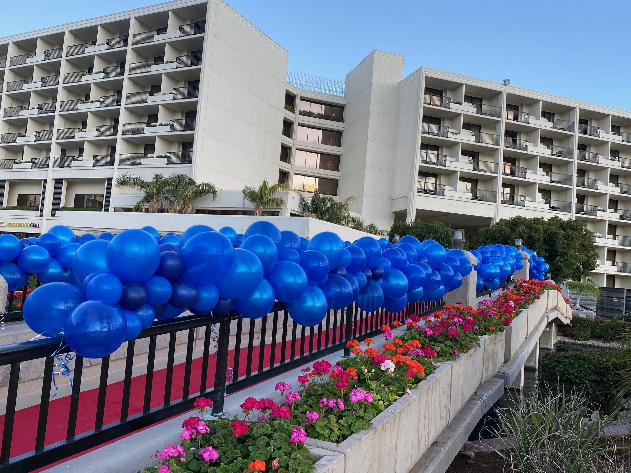 A multi-story white building with several balconies; a decorative blue balloon display lines a railing in front of the building; there are colorful flowers in a planter along the walkway.