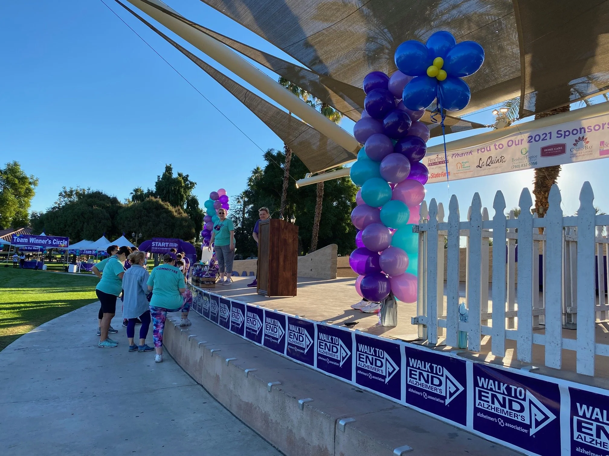 People participating in a Walk to End Alzheimer's event, standing behind a barrier with Alzheimer's awareness signs. The stage is decorated with purple and blue balloons, and a woman speaking at the podium under a shade structure.