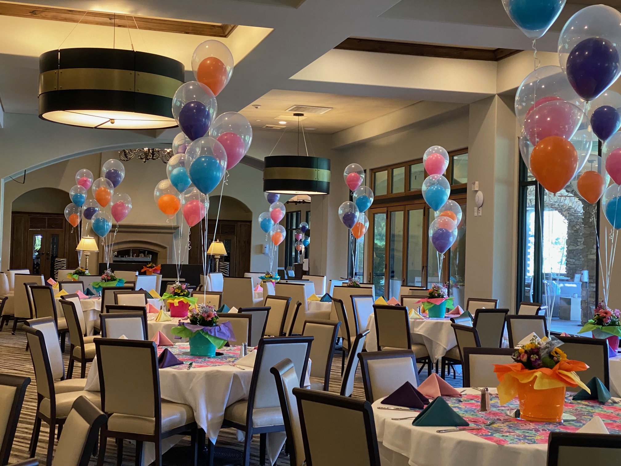 Decorated banquet hall with round tables covered in white tablecloths, colorful folded napkins, bordered by chairs with white cushions, and vibrant floral centerpieces. Balloons in various colors float above the tables, and the hall features large wi