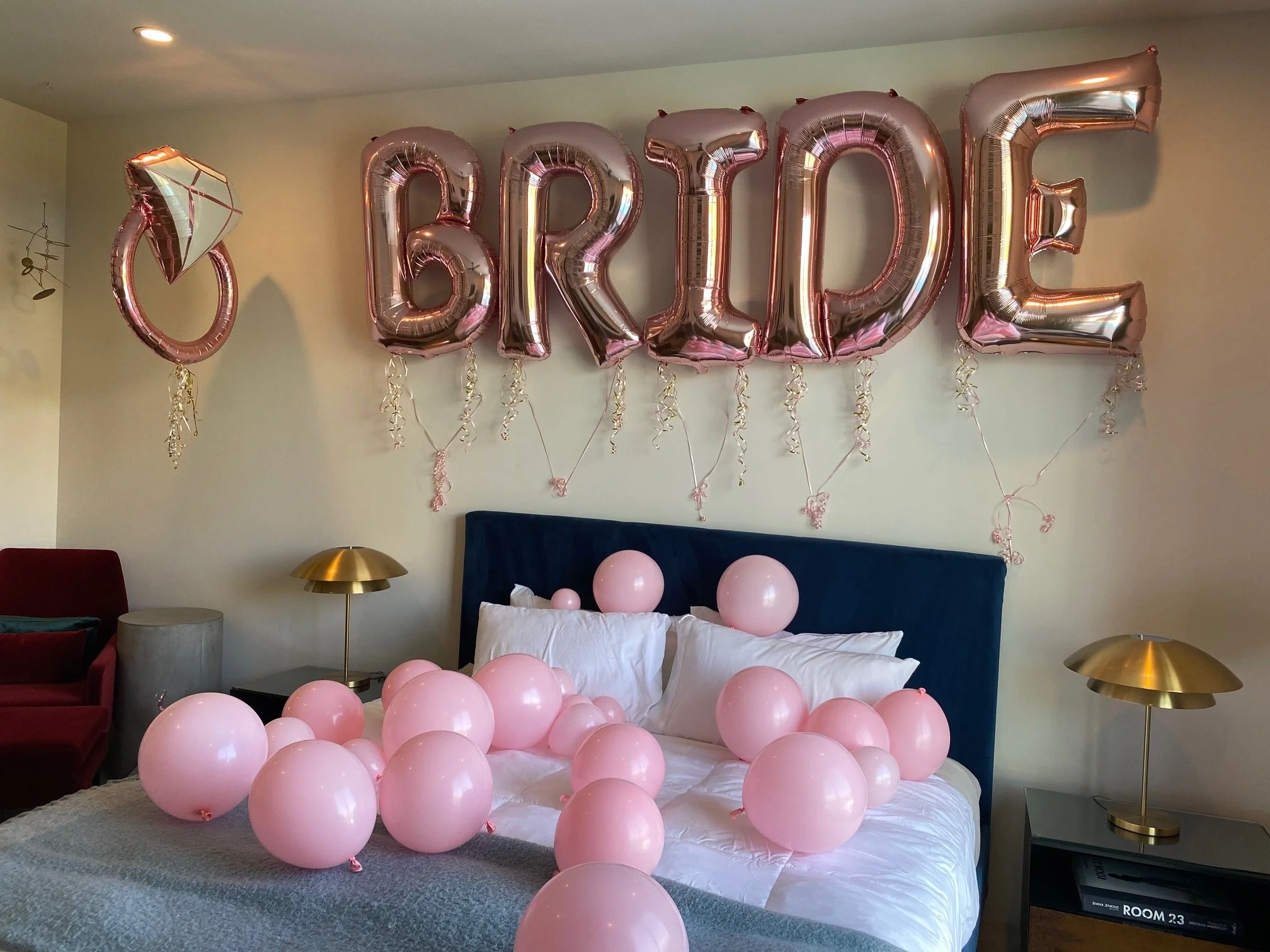 Room decorated with pink balloons and large foil balloons spelling 'BRIDE' on the wall, with pink balloon bouquets on the bed, and gold lamps on nightstands.