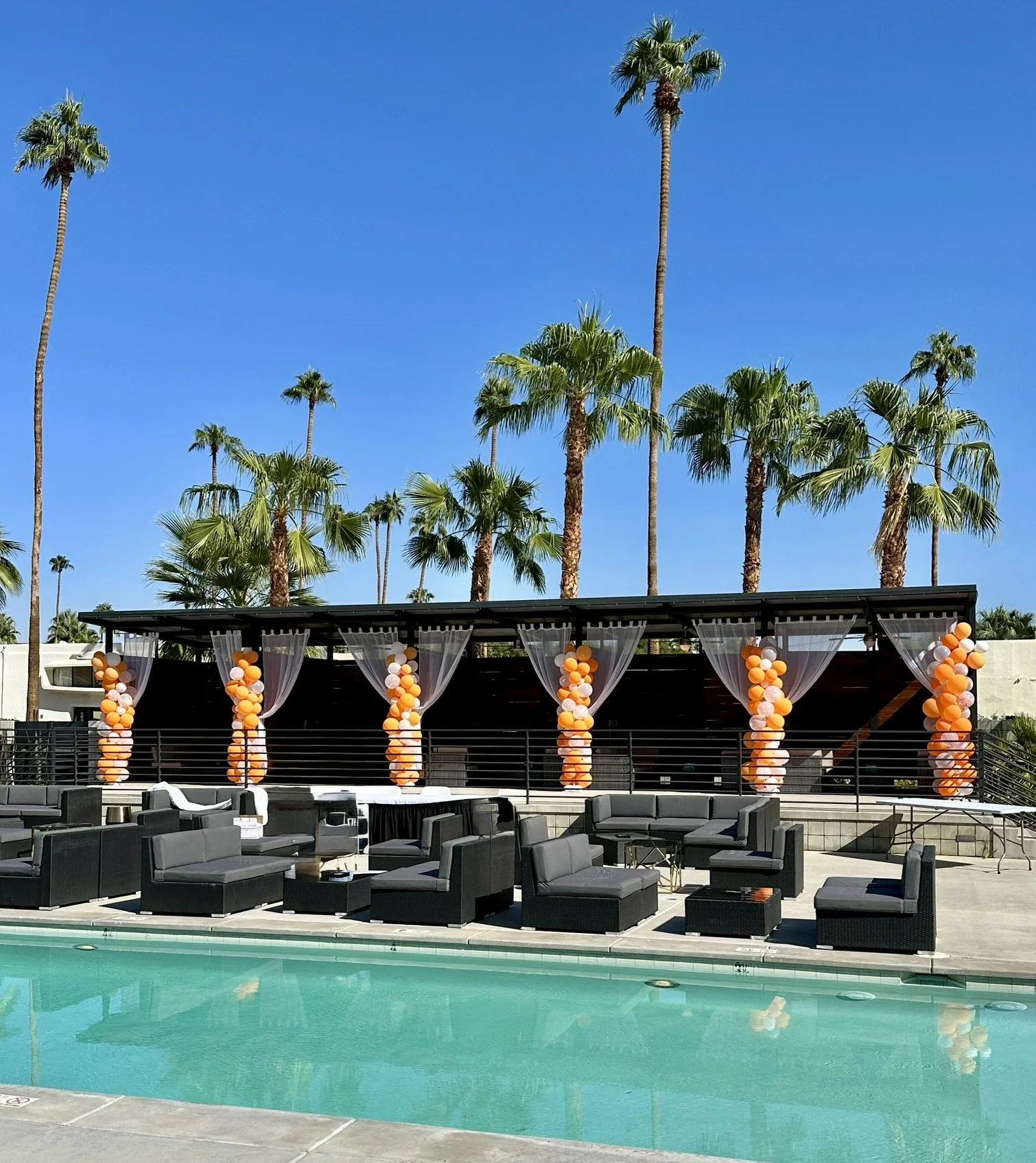 Poolside area with black outdoor furniture, decorated with orange and white balloons and draped curtains, against a backdrop of palm trees under a clear blue sky.