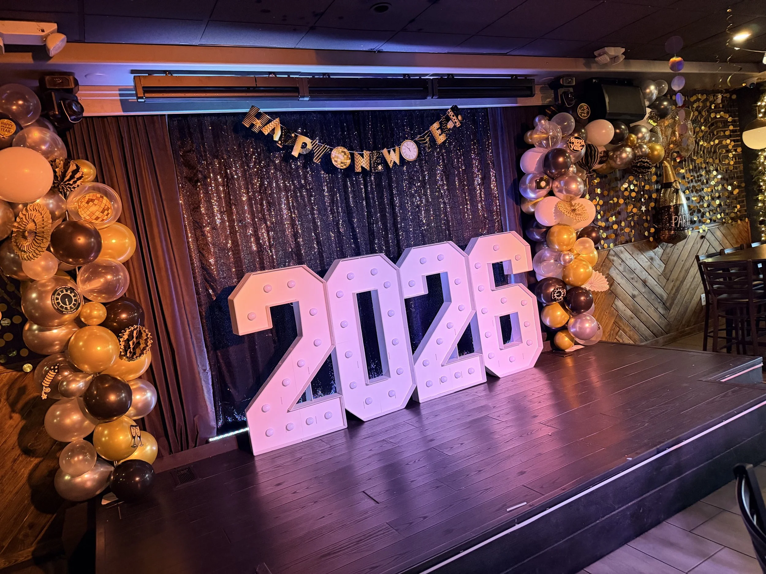 Decorations for New Year's celebration, including a large pink '2023' sign, black, gold, pink, and white balloons, and a glittery black curtain with a 'Happy New Year' banner and hanging clock.