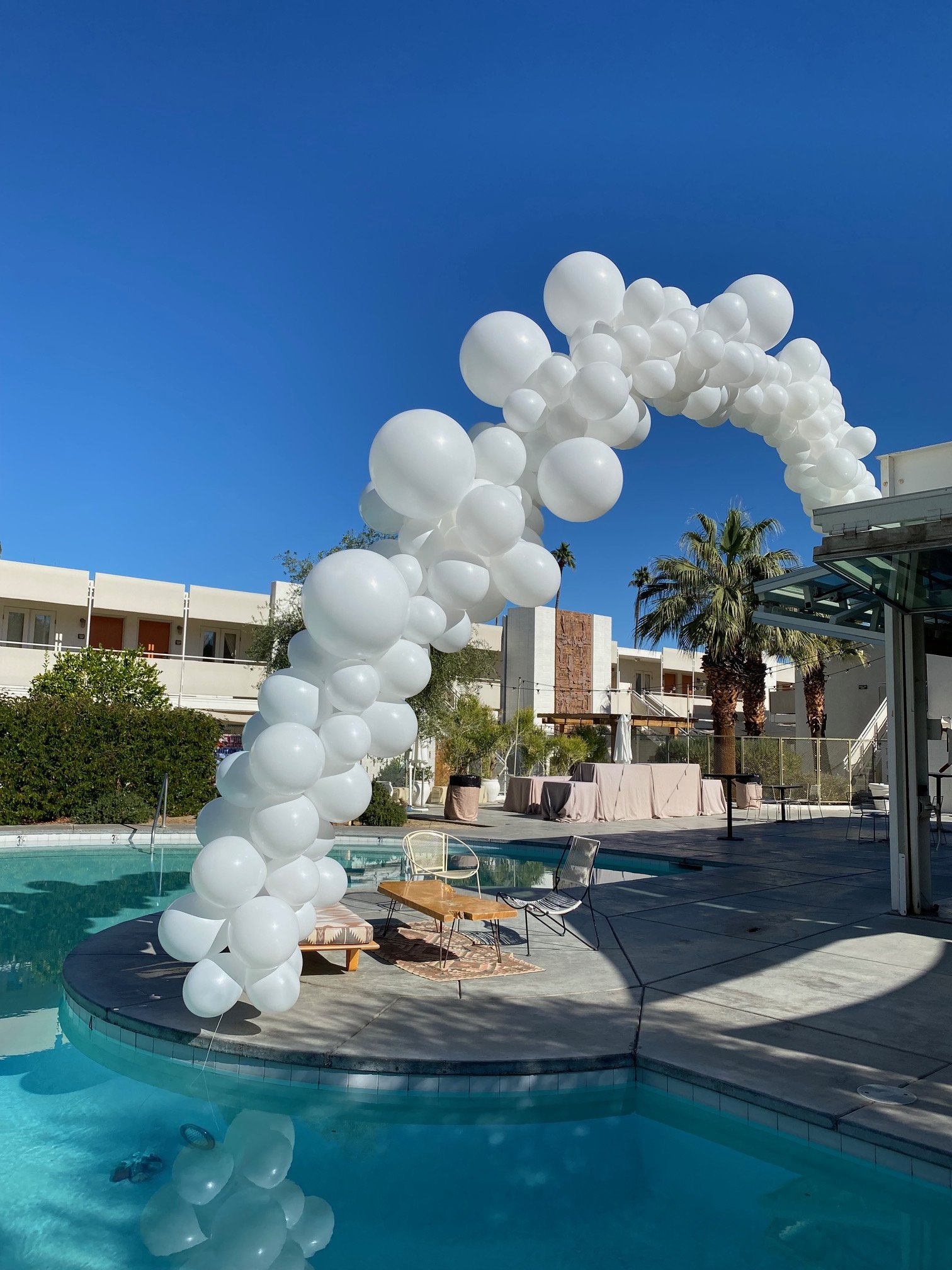 A poolside scene with a large balloon arch made of white balloons, outdoor lounge chairs, a table, palm trees, and modern buildings under a clear blue sky.