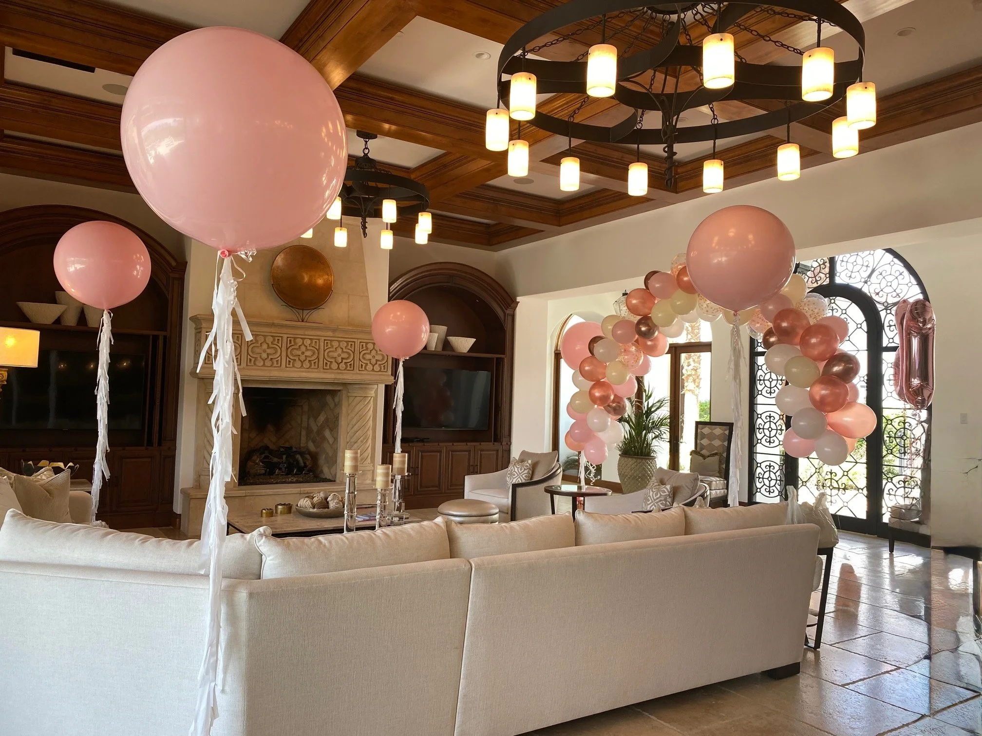 Living room decorated with pink, white, and copper balloons for a celebration, featuring a large sectional sofa, wooden ceiling with chandeliers, a fireplace, and large windows with decorative ironwork.