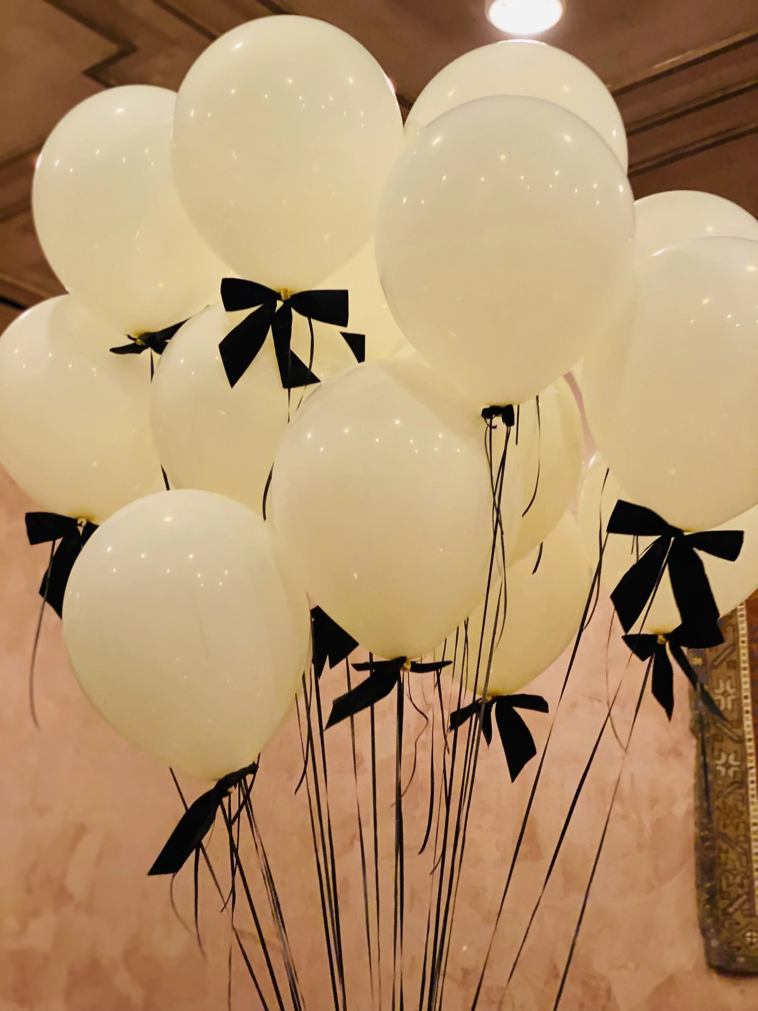 A bunch of white balloons with black ribbons tied around the stems, gathered together on a wooden floor in a room, with some lighting reflections visible on the balloons.