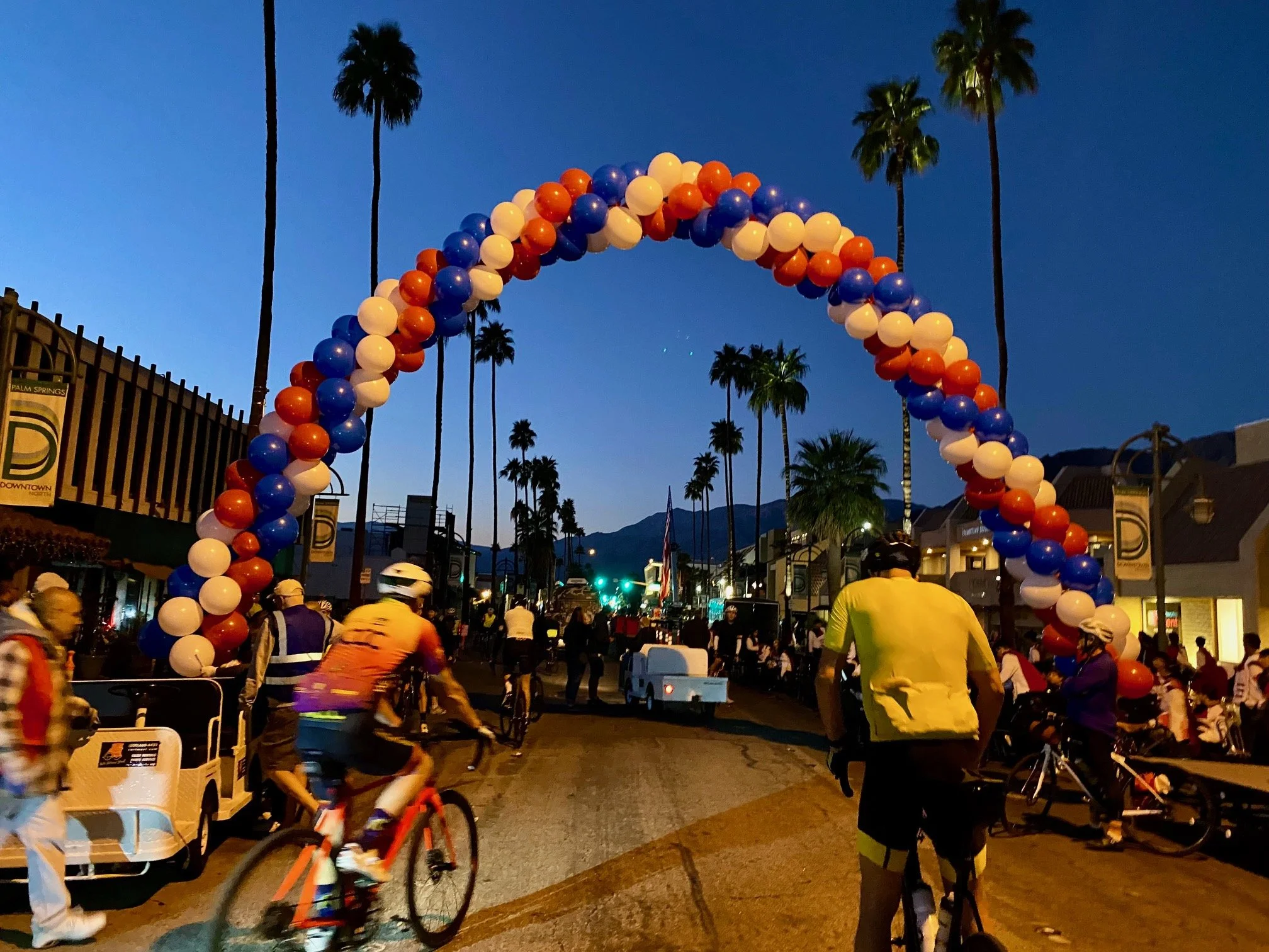 Red, white and blue balloon arch over a street with cyclist underneath it. 