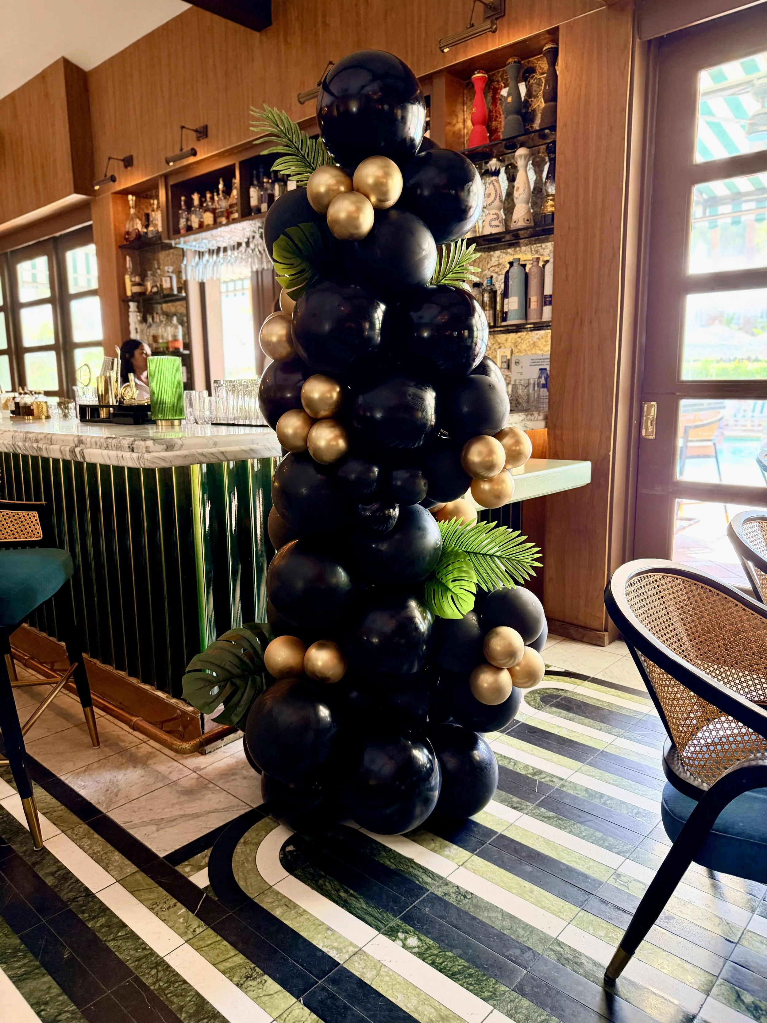 Decorative column made of black, gold, and beige balloons with green leaves in a restaurant or bar setting, featuring a marble countertop, glassware, and wooden shelves with bottles and decorations.