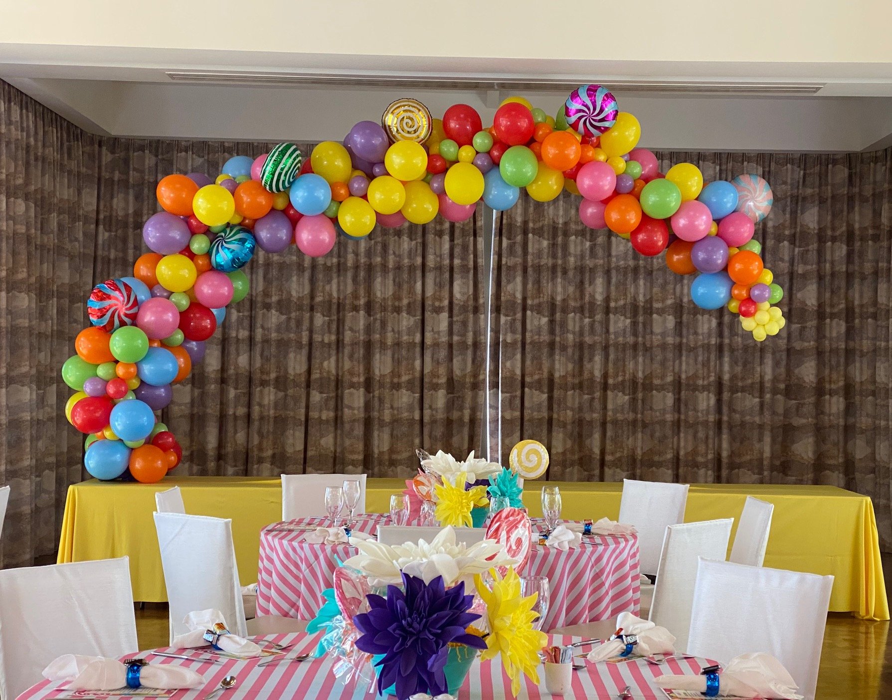 Colorful balloon arch with yellow, red, purple, green, orange, blue, and striped balloons decorating a party hall with tables set for a celebration.