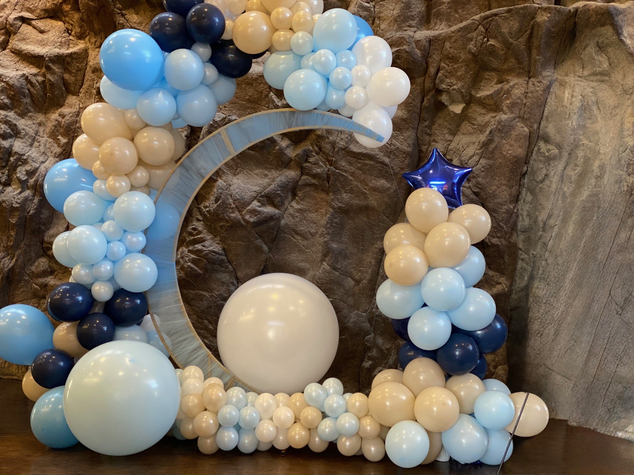 Decorative balloon arrangement with blue, white, cream, beige, black, and a blue star-shaped balloon against a stone wall background.