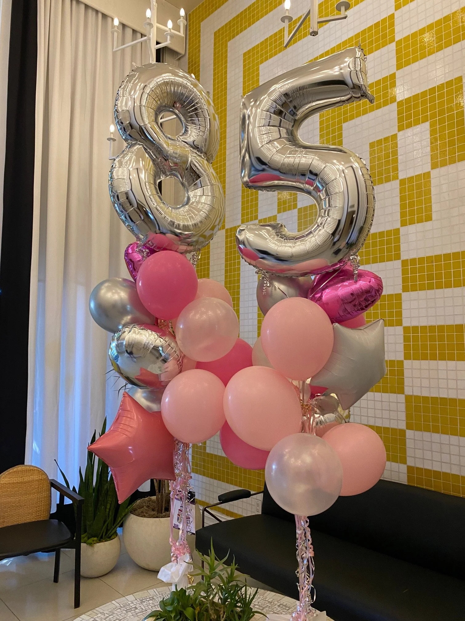 Decorative balloons, including silver number balloons forming '85' and pink, silver, and heart-shaped balloons, arranged for a celebration inside a room with a yellow and white tiled wall and potted plants.