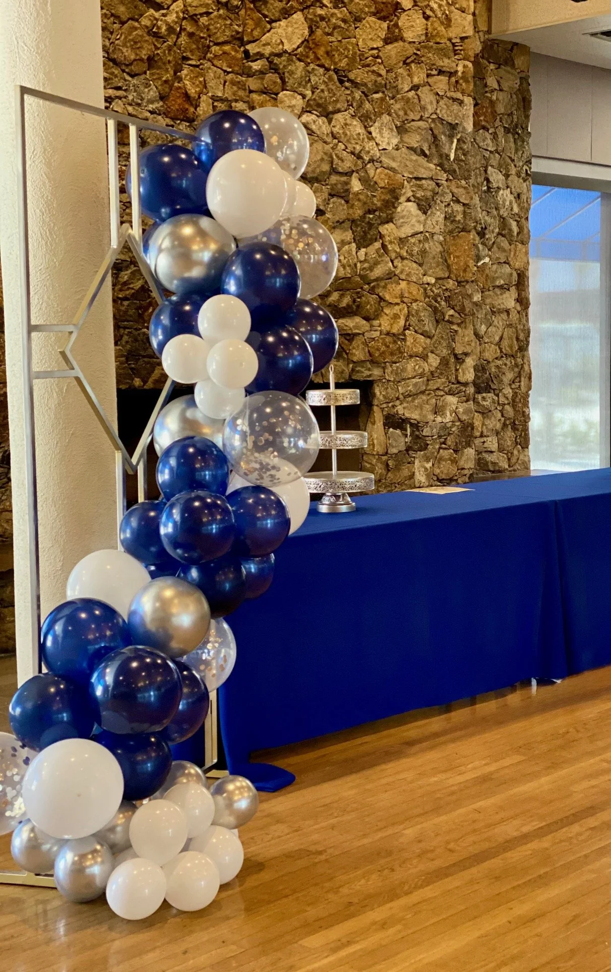 A balloon decoration with white, navy blue, clear balloons, and metallic balloons with confetti is arranged next to a blue-draped table with a three-tiered silver tray on top, set against a stone wall in an indoor setting.