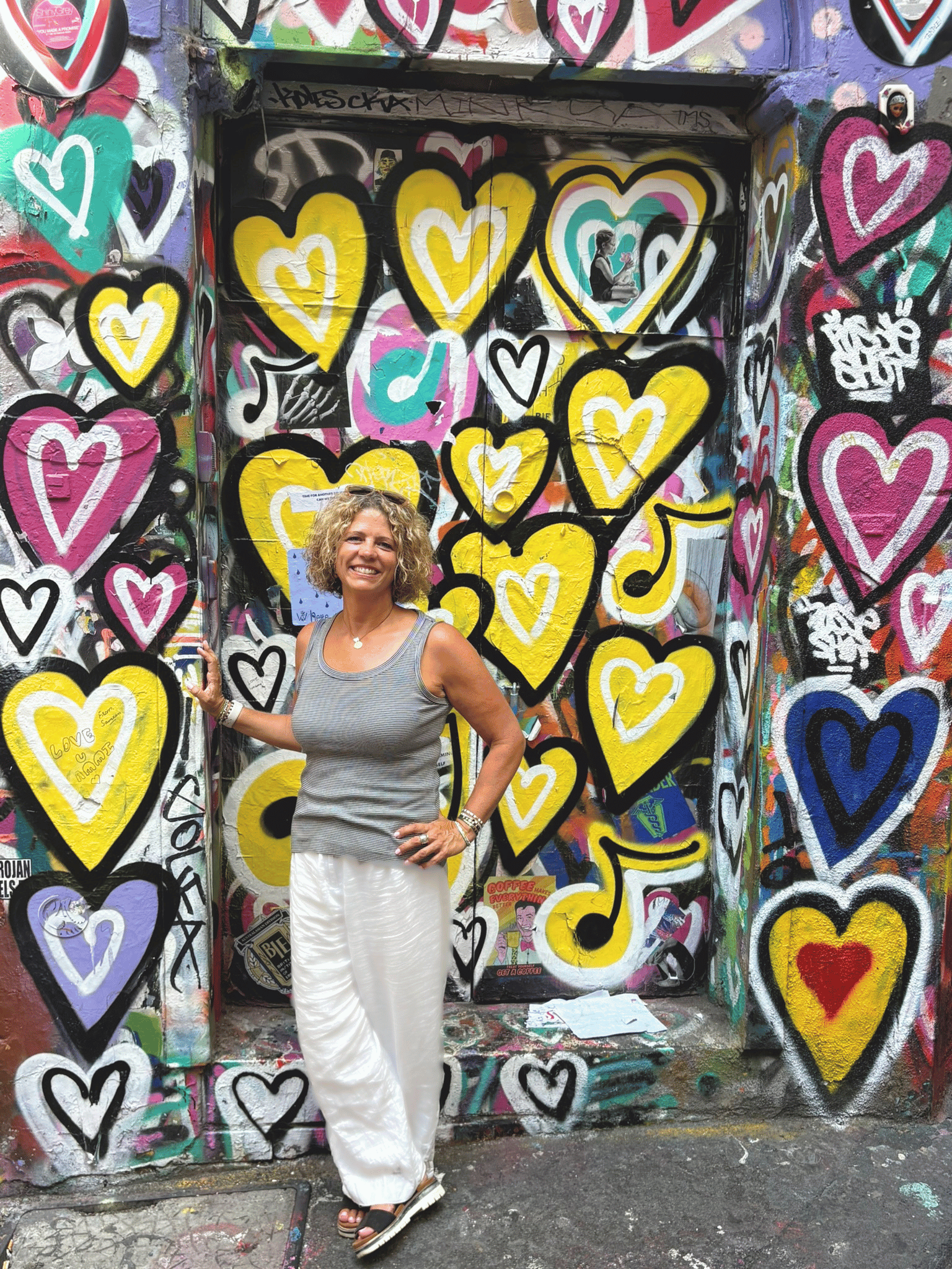 A woman standing in front of a wall covered in colorful graffiti featuring numerous hearts, with some containing smaller hearts or designs. The woman is smiling, wearing white pants, a gray sleeveless top, and sandals. She has curly blonde hair and sunglasses resting on her head.