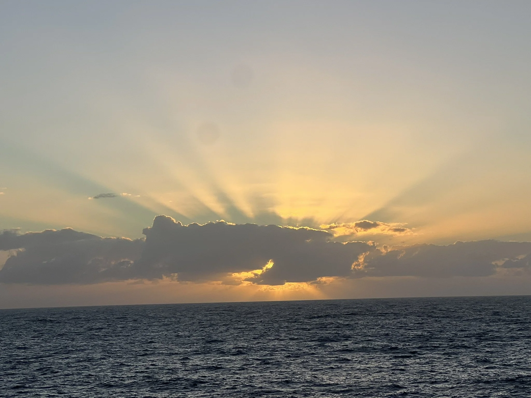 Sunset over the ocean with dark clouds near the horizon and sun rays shining through the clouds.