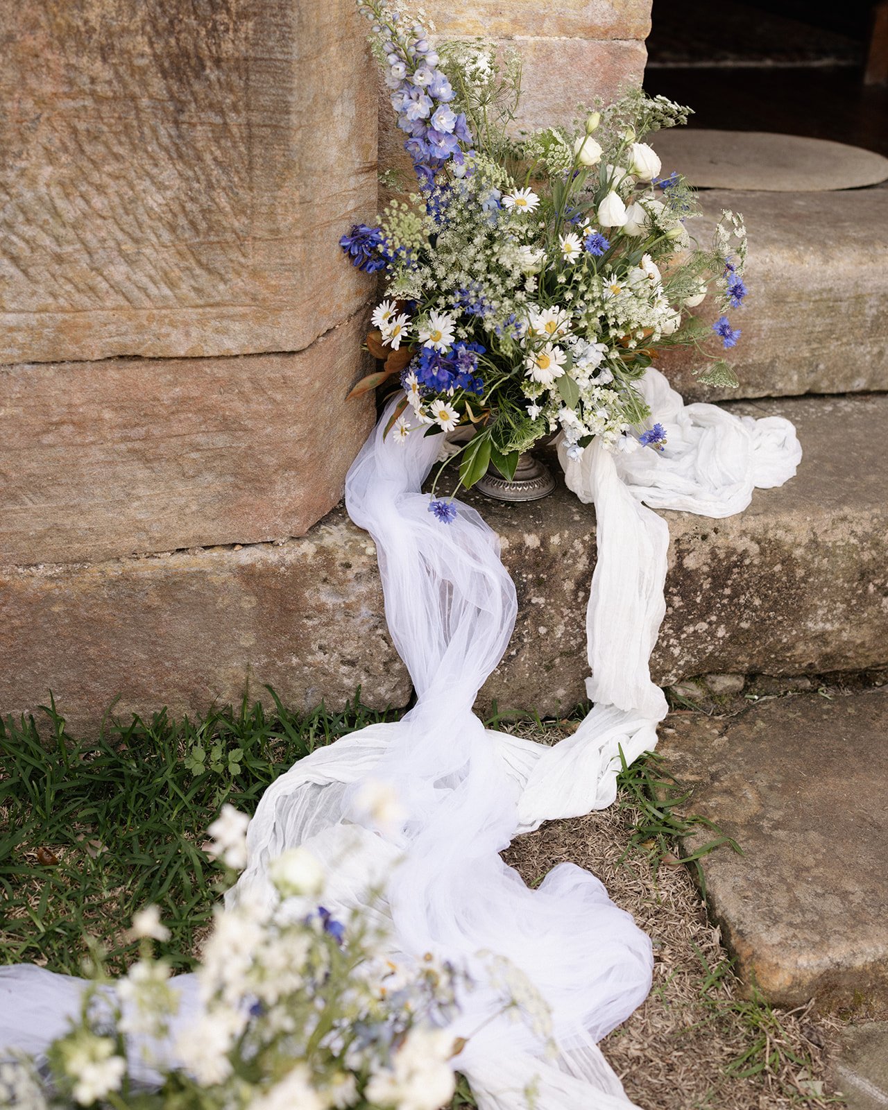 A bouquet of white, purple, and blue flowers arranged on a stone step, with flowing white fabric draped around the flowers at a wedding conducted by marriage celebrant Kate Cohen outside of Sydney in Wisemans Ferry