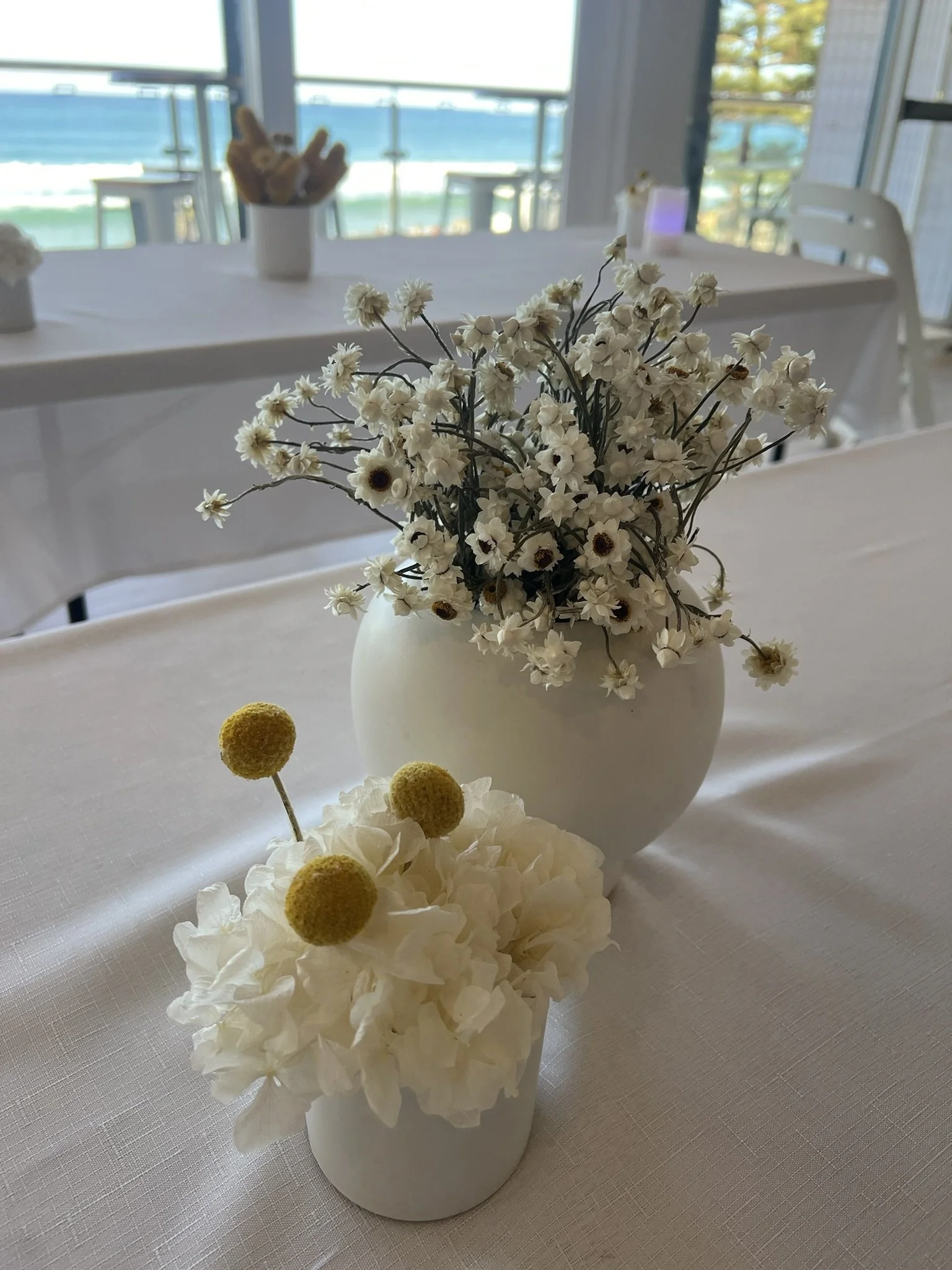 A white vase filled with small white flowers with cream and brown blossom centers, set on a white tablecloth at a funeral in a surf lifesaving club on Sydney's northern beaches for a funeral