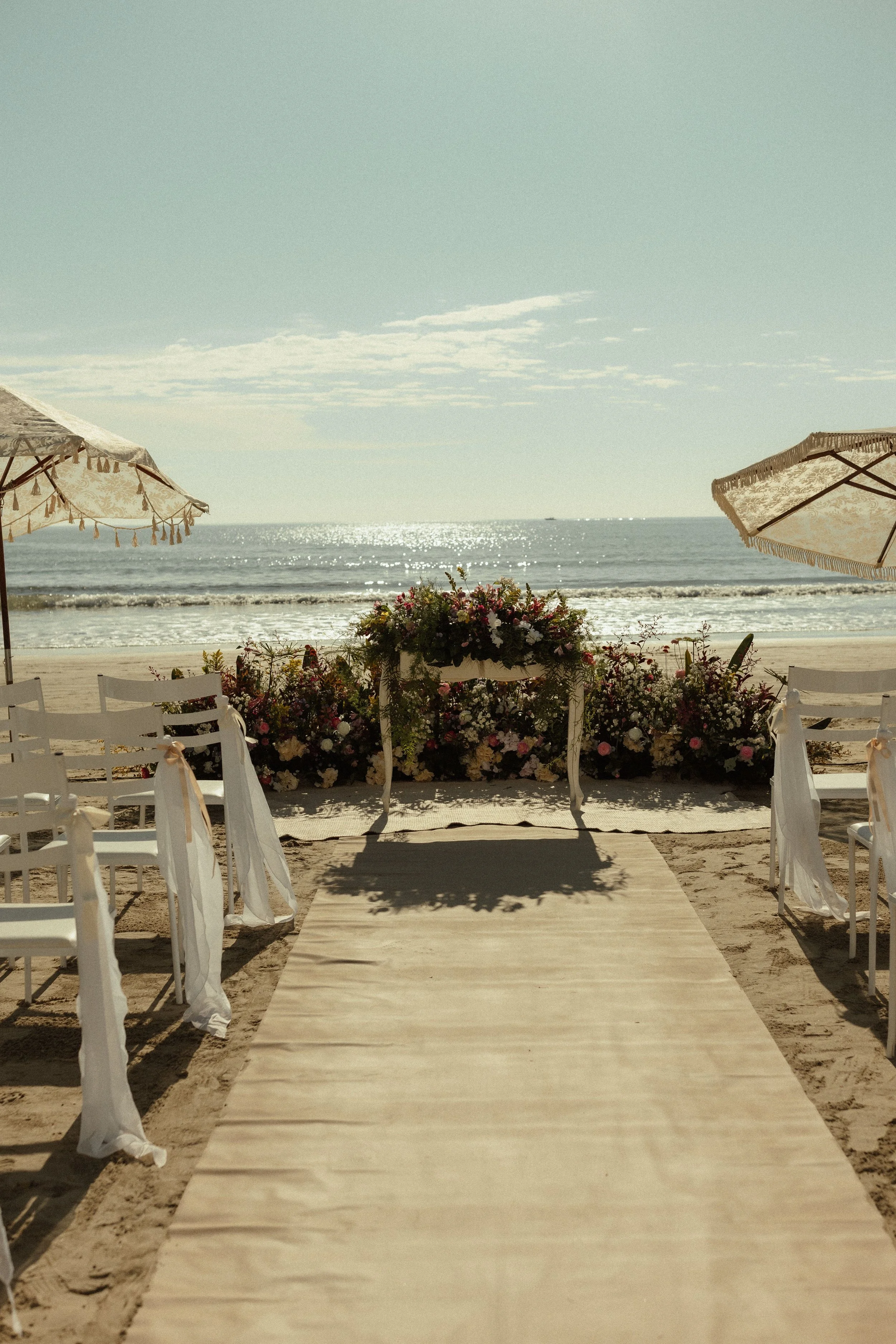 A beach wedding setup with a aisle runner, white chairs with fabric sashes, floral arrangements, and umbrellas with tassels, overlooking the ocean on a sunny day.