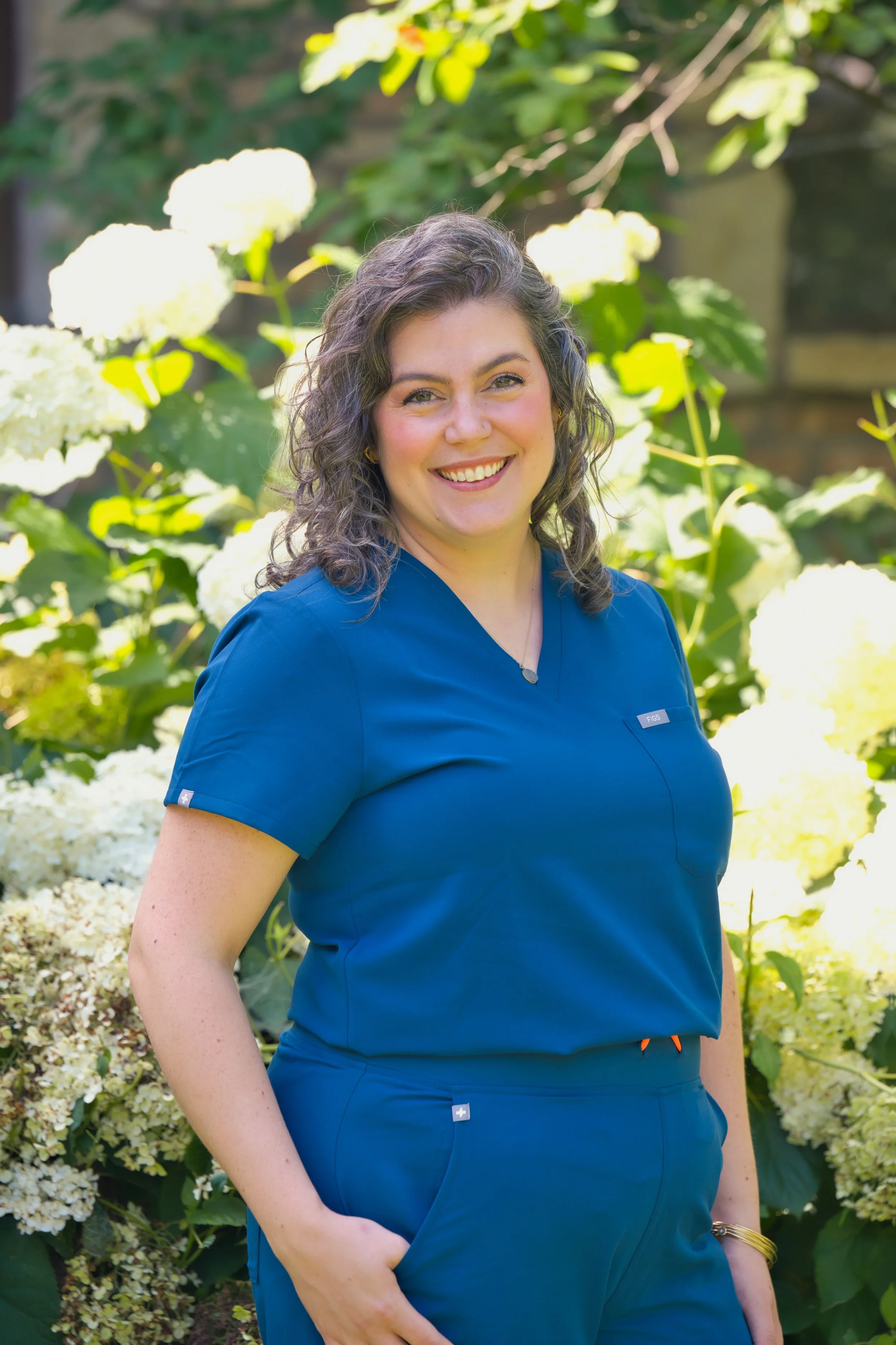 A smiling woman with curly hair and a nose piercing, wearing blue medical scrubs, standing outdoors with white hydrangea flowers and green foliage in the background.
