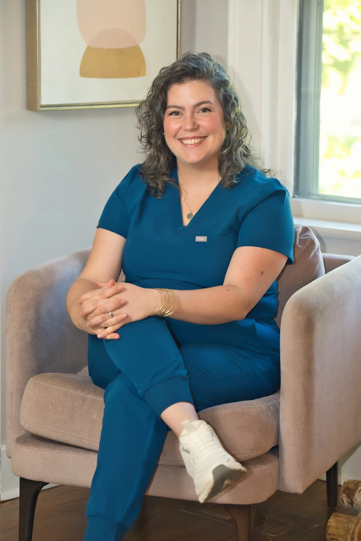 A woman with curly gray hair, wearing blue medical scrubs, sitting on a beige armchair by a window with a smile, with her hands clasped on her lap and her legs crossed. A framed artwork hangs on the wall behind her.