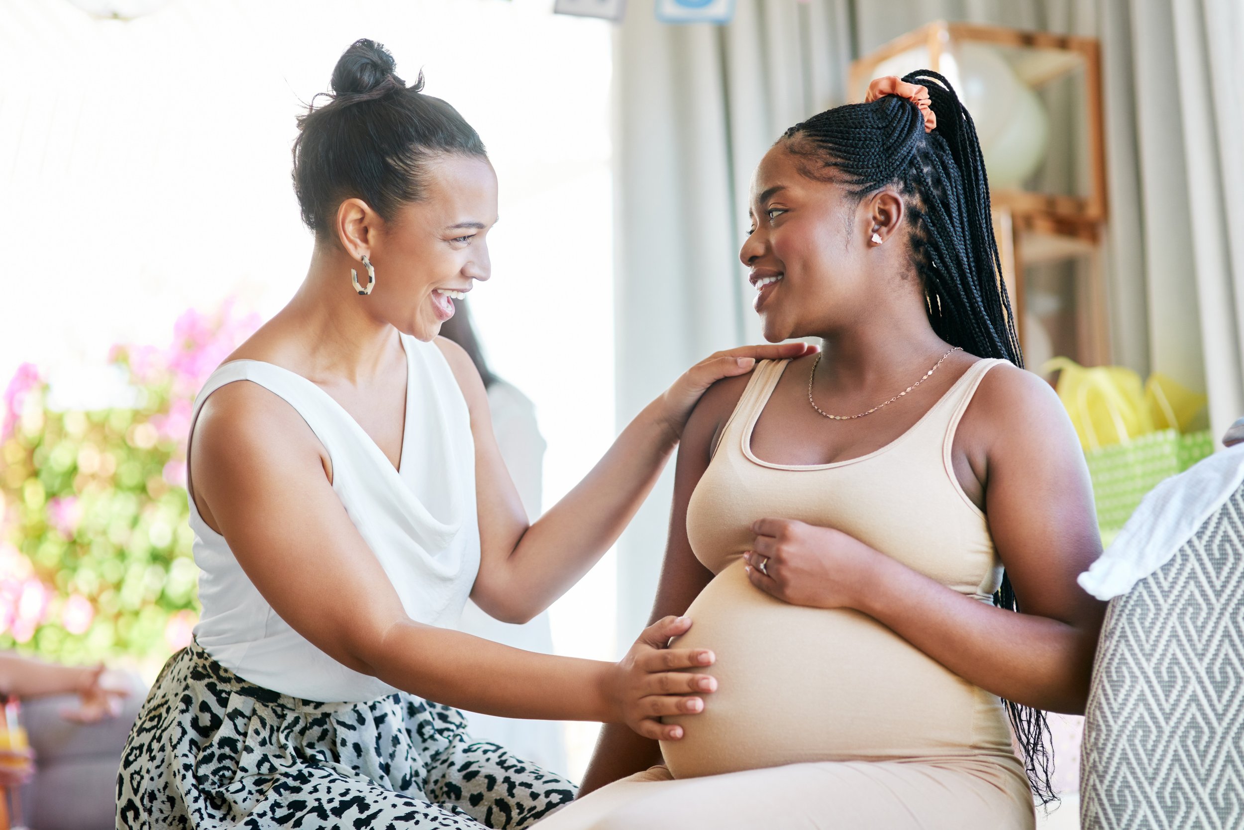 Pregnant woman smiling as she interacts with a woman in a white top and leopard-print skirt, inside a bright room with a blurred background of plants and furniture.