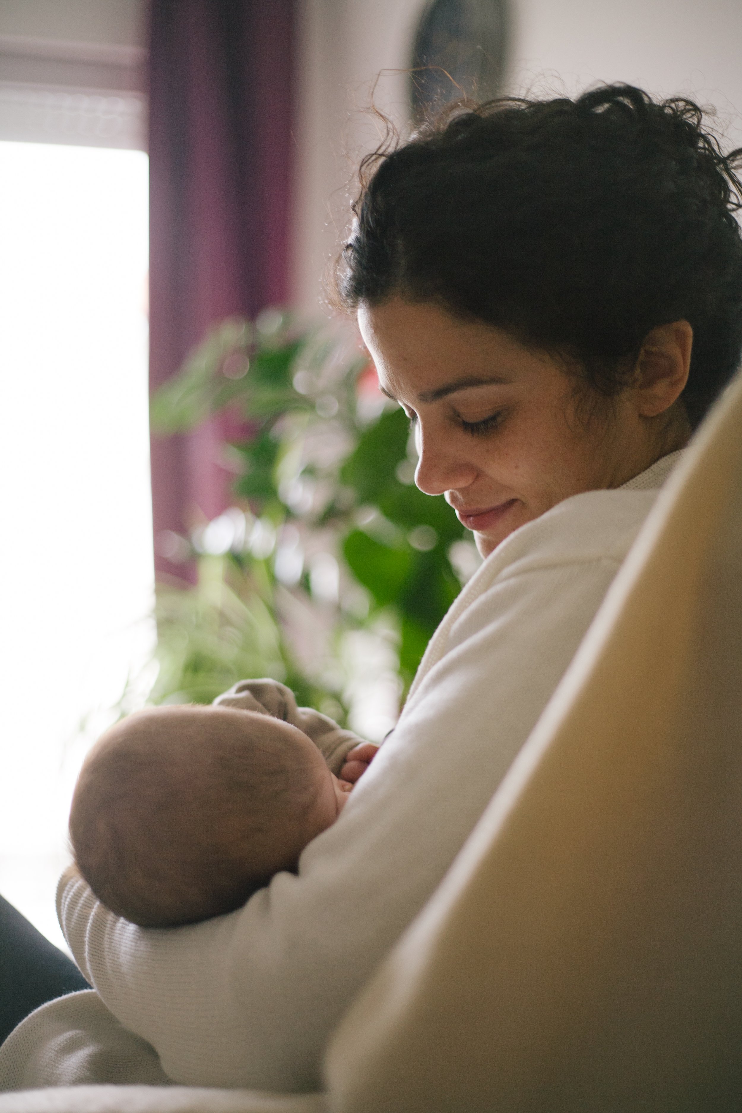 A young woman with curly dark hair and freckles holding and smiling at a newborn baby indoors near a window with a blurred purple curtain and green plants in the background.