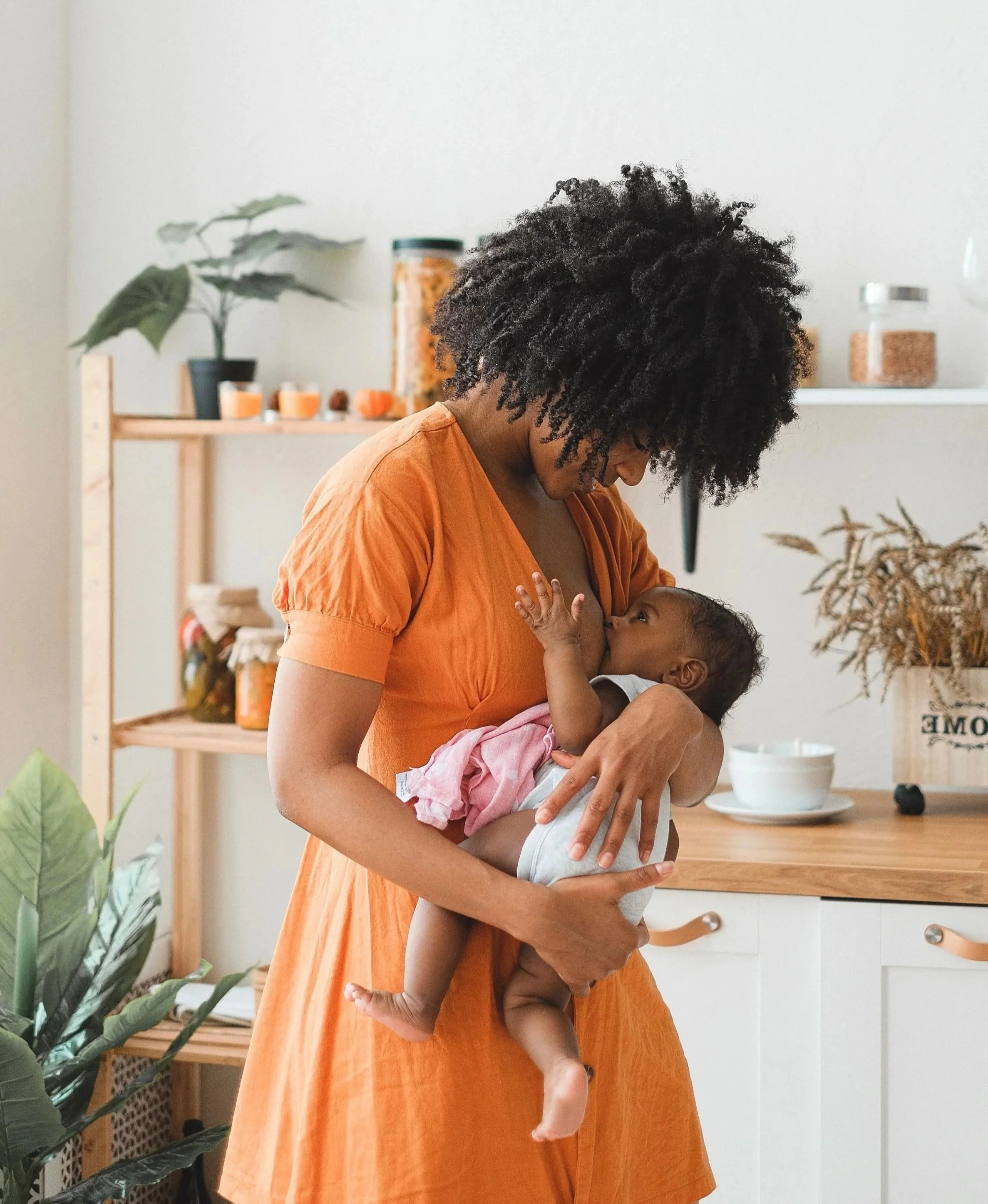 A woman with curly black hair wearing an orange dress holding a young child with dark skin and curly hair in a kitchen. The woman is nursing the child. There are houseplants and kitchen items in the background.