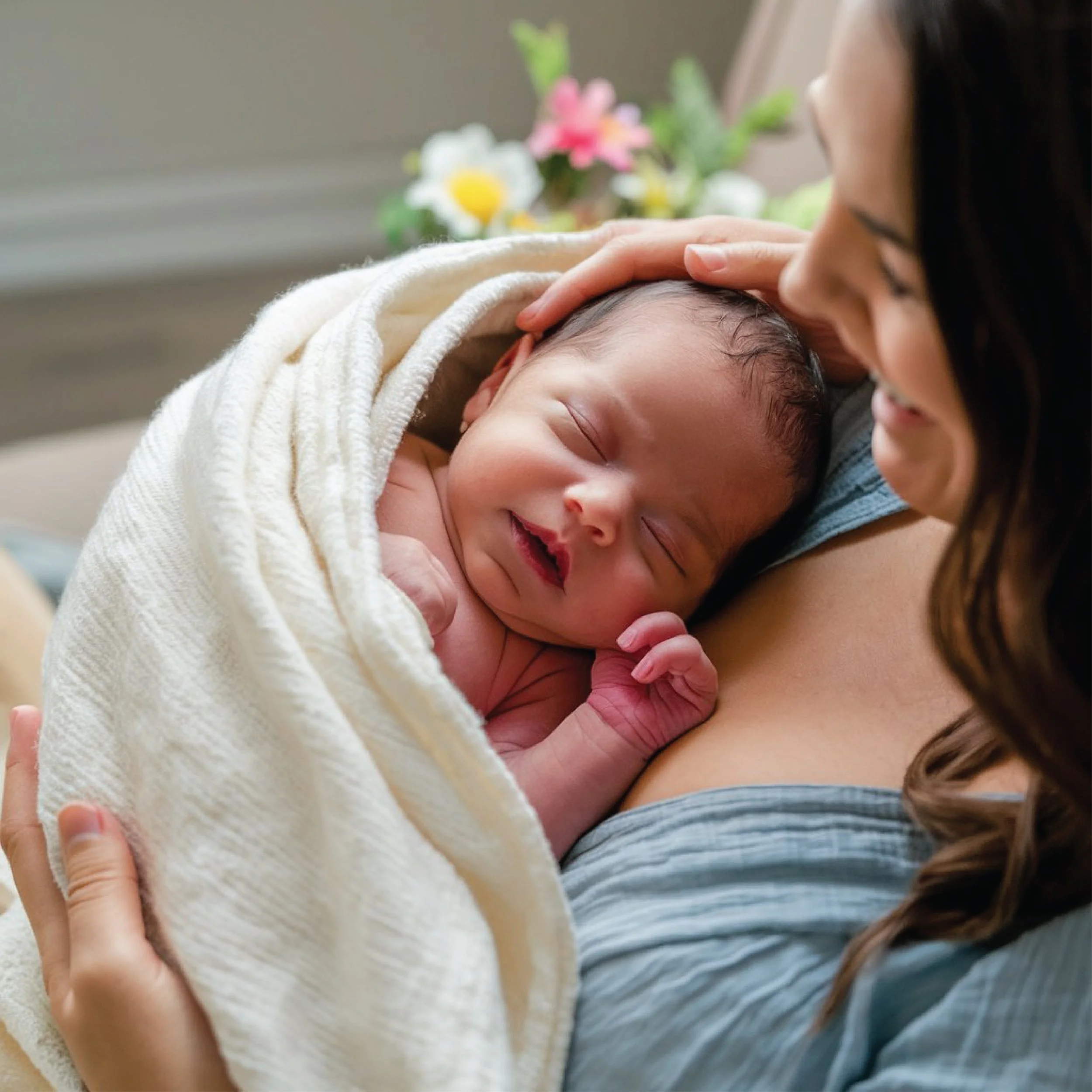 A woman with long brown hair holding a sleeping baby, who has a small smile and is wrapped in a white blanket, with blurred flowers in the background.