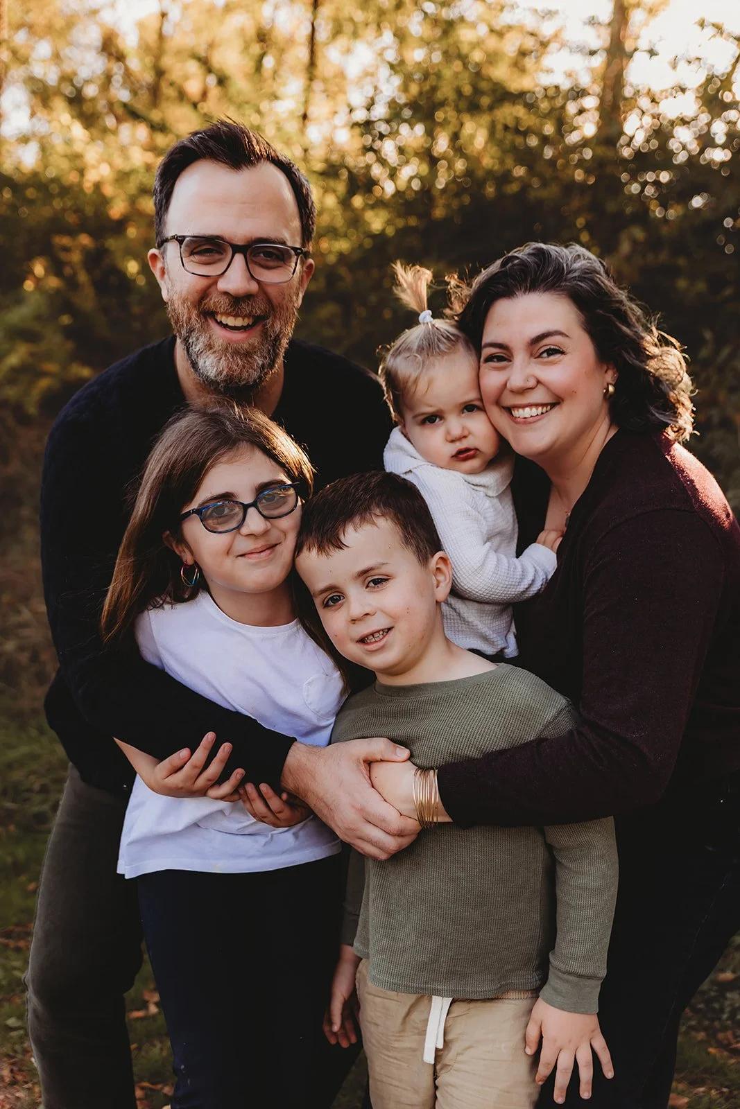 A family of six smiling and embracing outdoors during golden hour with trees in the background.