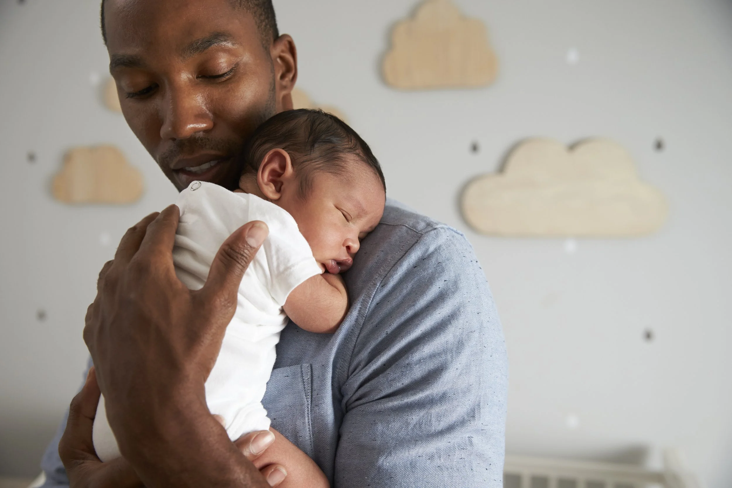 A man lovingly holding a sleeping baby close to his chest indoors, with a background of a wall decorated with cloud-shaped patterns.