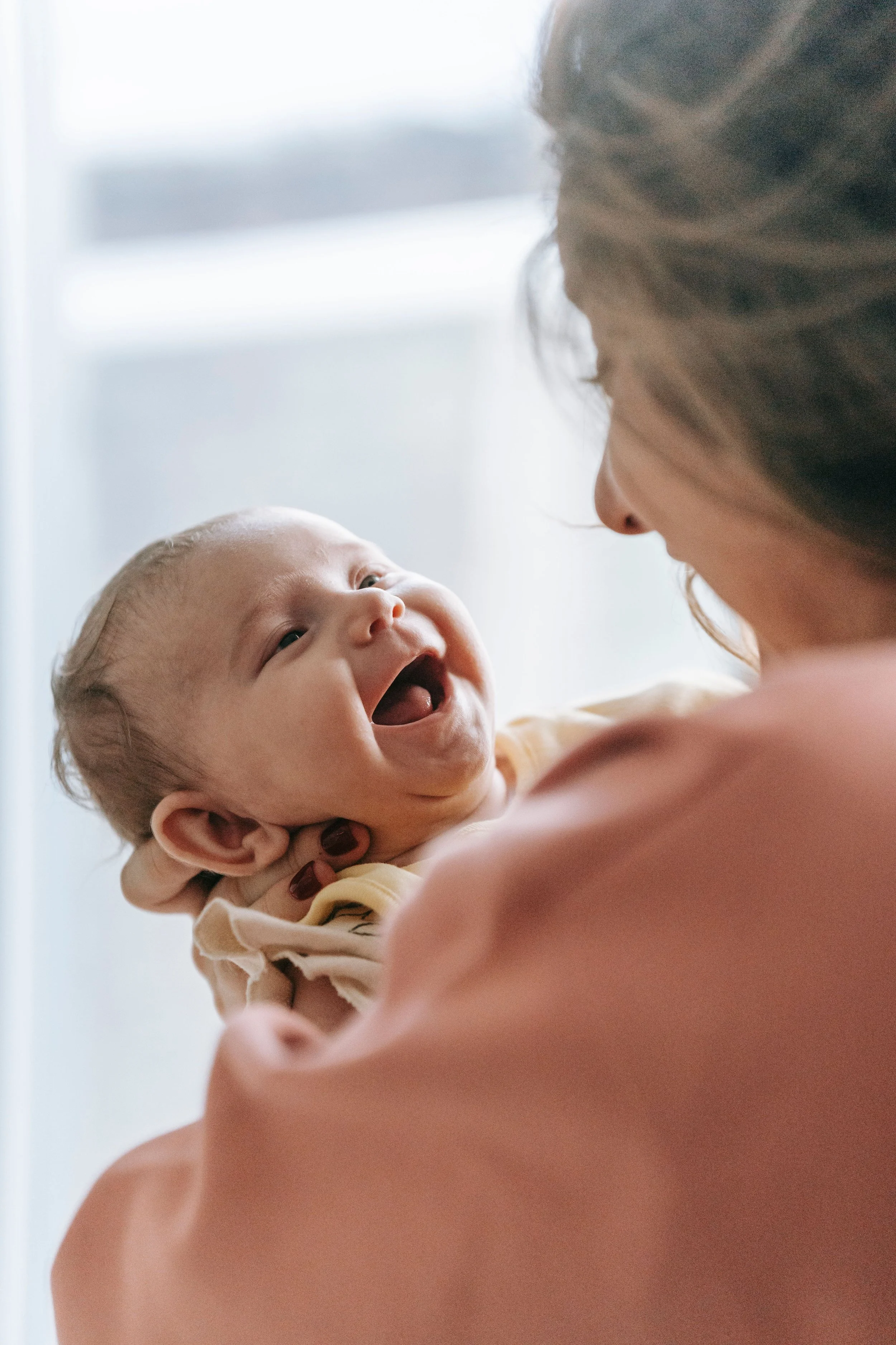 A mother lifting her smiling baby, looking into each other's eyes, with soft natural light background.