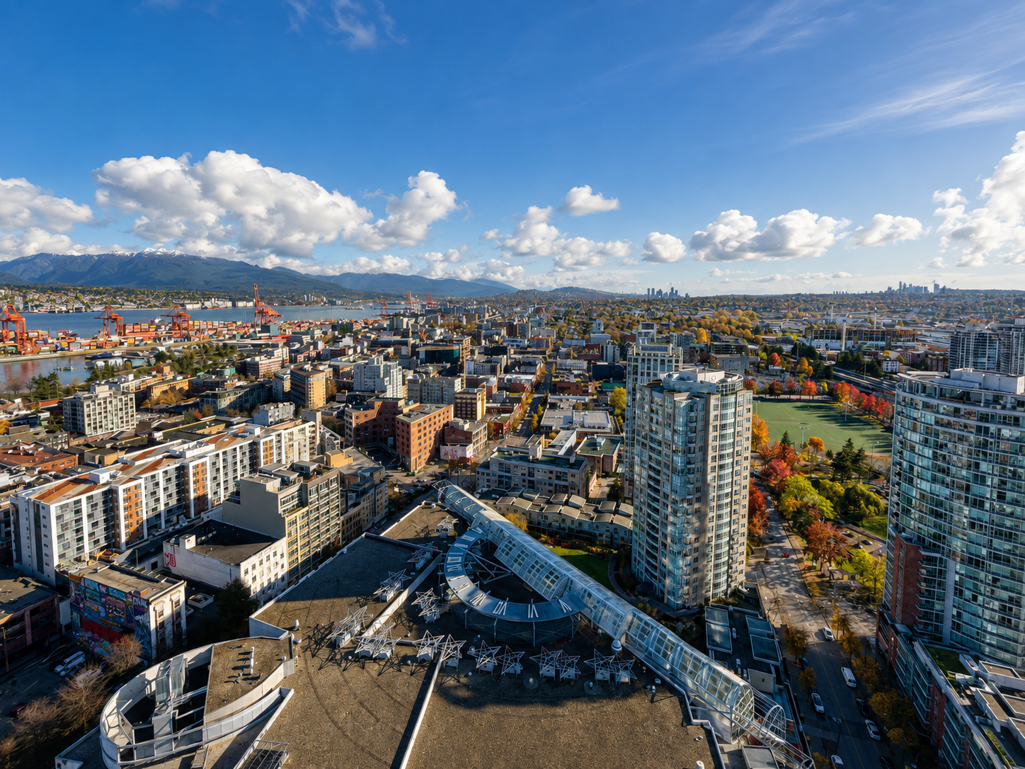 Aerial view of a cityscape with tall buildings, a river with cranes, and mountains in the background under a partly cloudy sky during daytime.