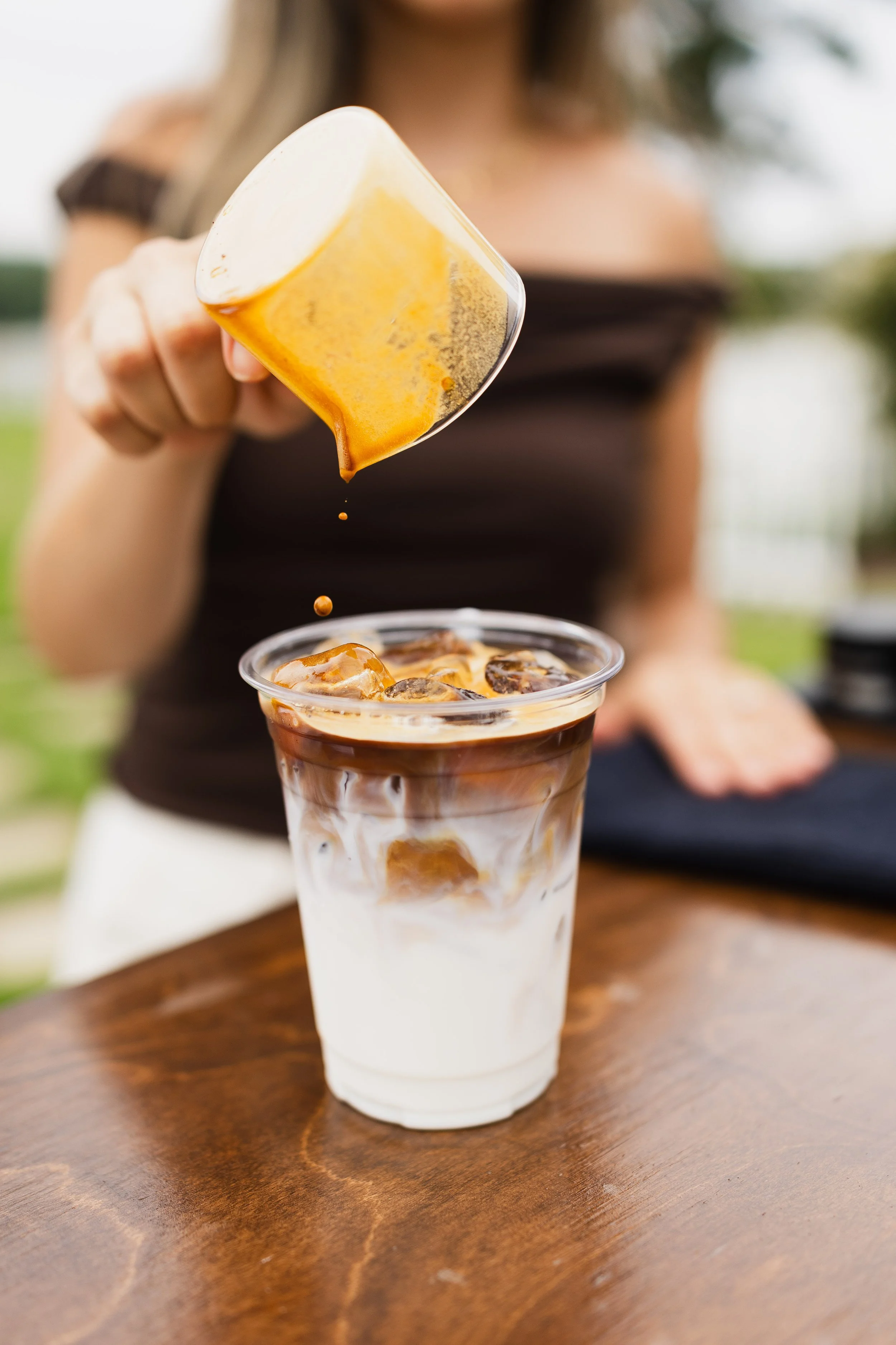 A woman is holding a small cup of caramel sauce over a glass of iced coffee with milk and ice cubes on a wooden table, outdoors.