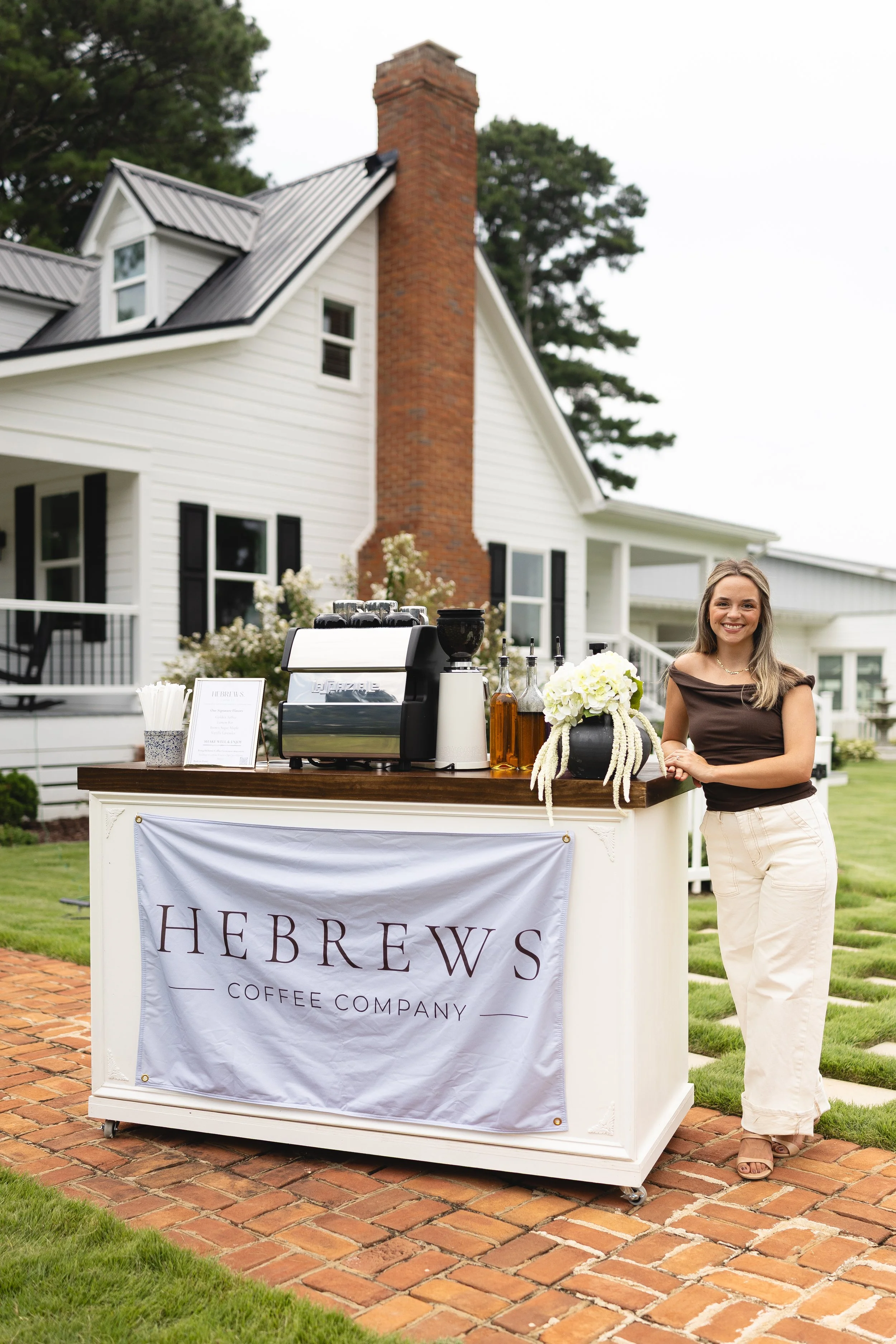 A woman standing next to a coffee stand with a sign that reads 'Herbrew Coffee Company' in front of a white house with black shutters and a brick chimney, on a brick pathway with a lawn and trees in the background.