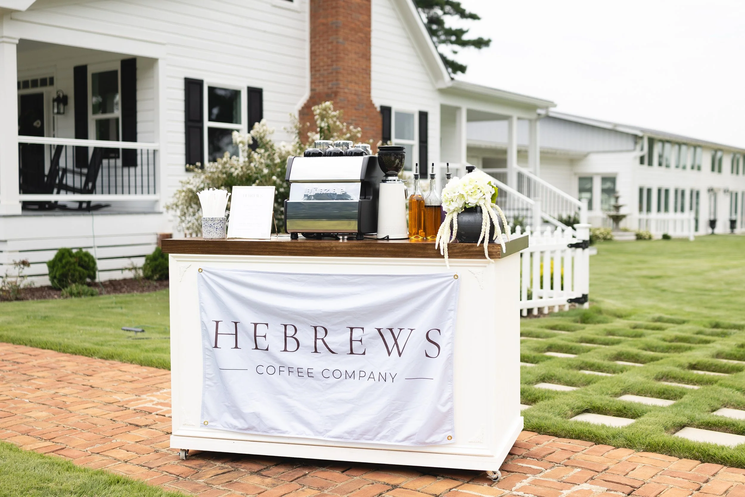Outdoor beverage station set up on a table with a sign reading 'Herb E W Coffee Company' in front of a white house with black shutters and a brick chimney, surrounded by a garden and lawn.