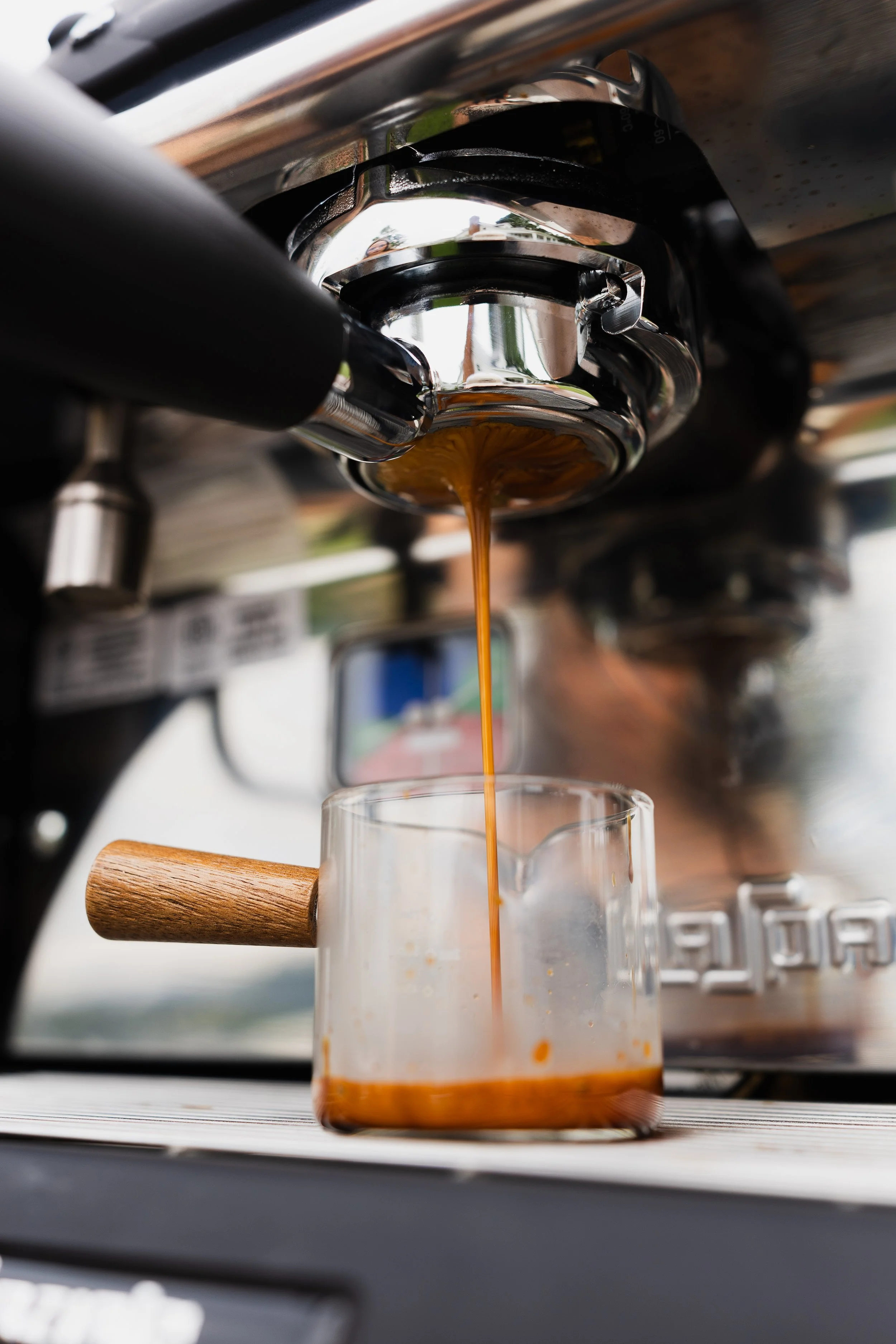 Close-up of a coffee machine pouring espresso into a clear glass mug, with a wooden handle on the mug and a blurred background.