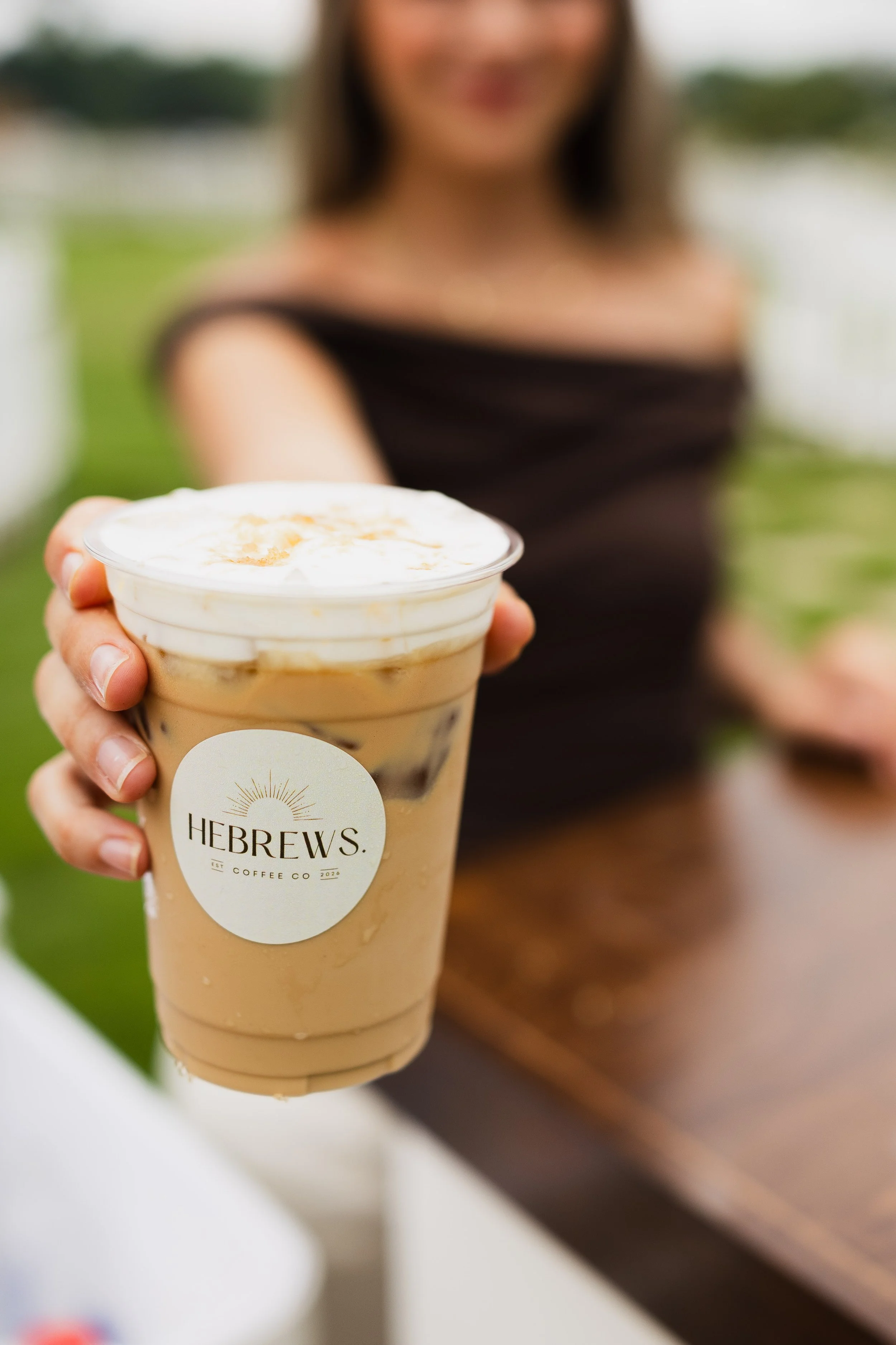 A woman holding a cold coffee drink in a clear plastic cup labeled 'Hebrews Coffee Co.' with a foamy top and caramel drizzle, outdoors with blurred green background.