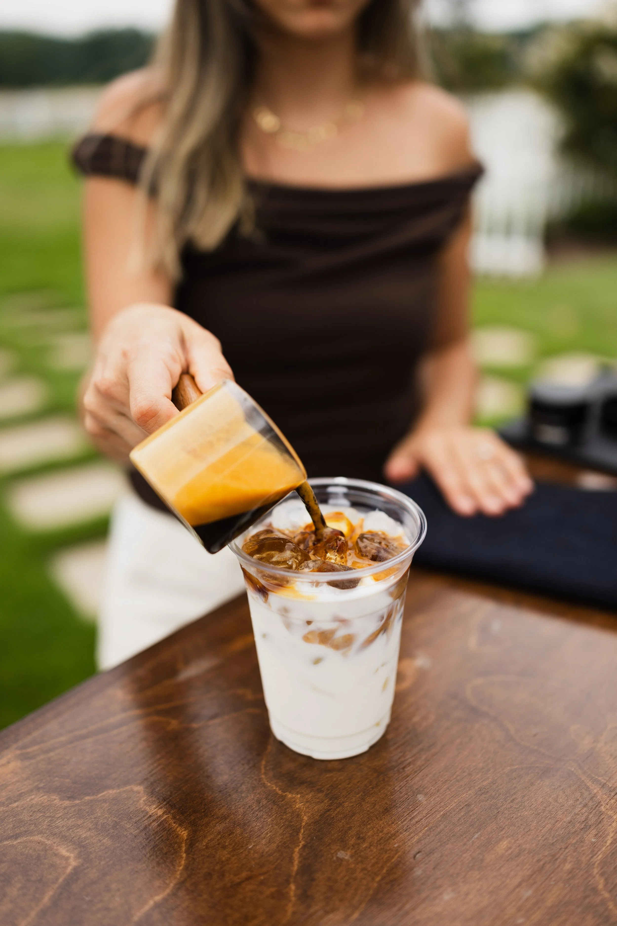 A woman in a black off-shoulder dress pouring coffee into a tall glass of iced coffee with whipped cream and caramel syrup, on a wooden table outdoors.