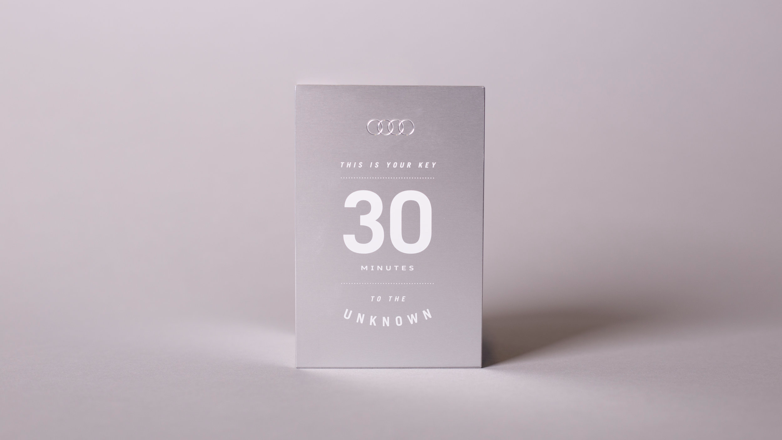 A minimalist gray card with the Audi logo at the top, and the text 'THIS IS YOUR KEY 30 MINUTES TO THE UNKNOWN' printed in white.