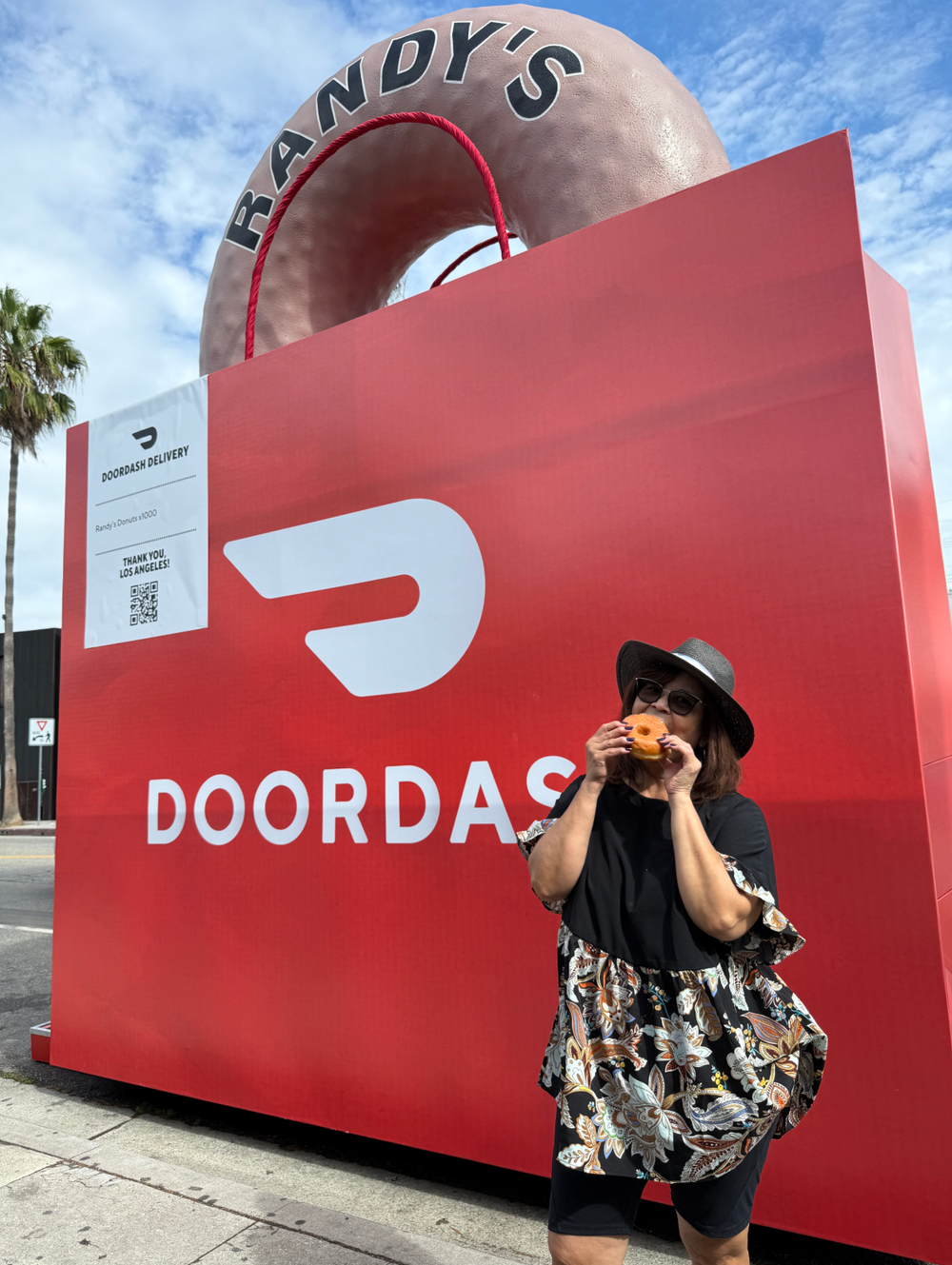 Woman wearing a black dress, hat, and sunglasses eating a donut in front of a large red DOORDASH sign with a giant donut structure on top. Palm trees and a partly cloudy sky are visible in the background.