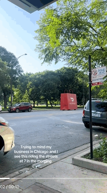 A parking lot with cars, trees, and a large red shopping bag on the ground. A sidewalk with a pole and a sign is in the foreground. The sky is clear with some clouds.